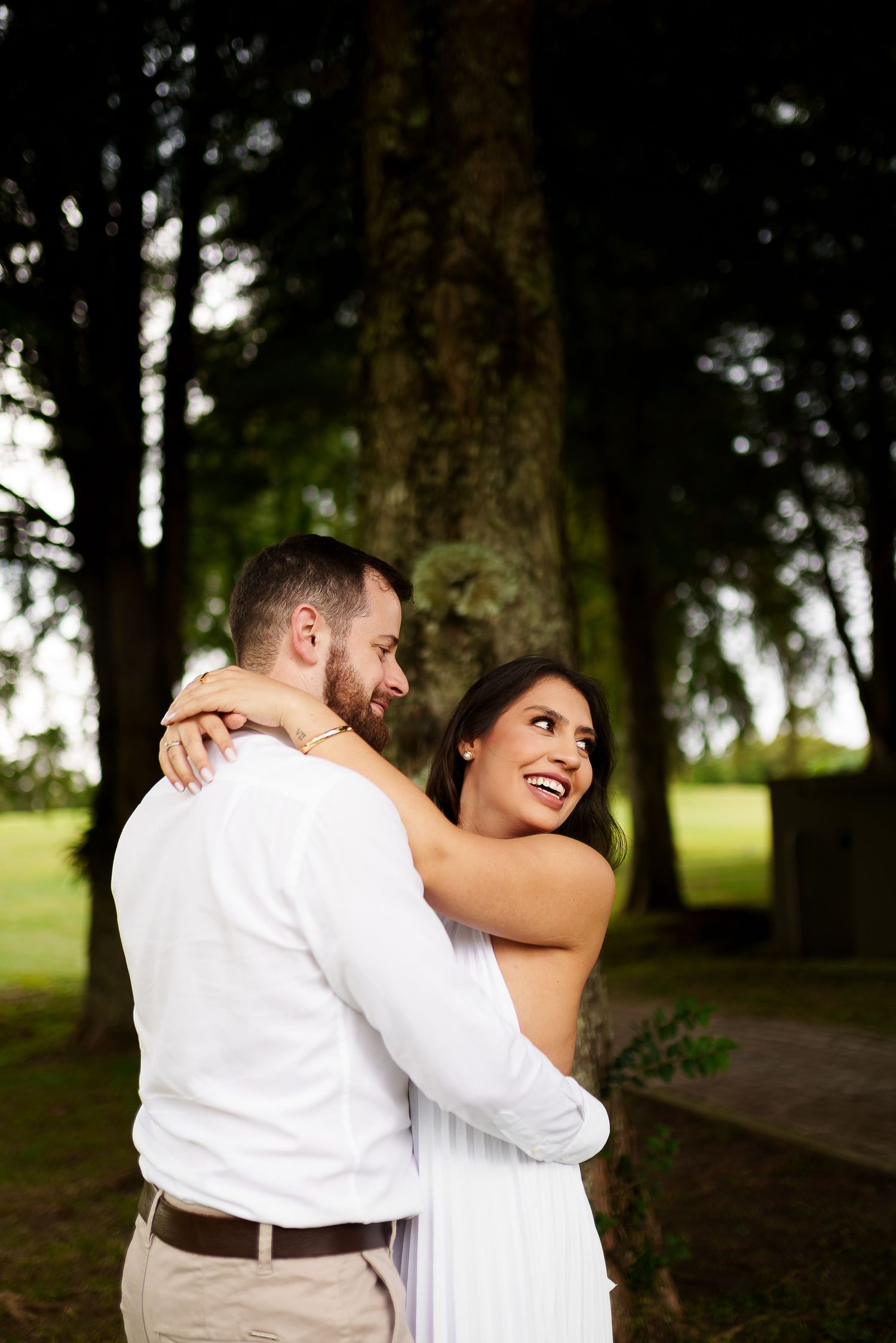 A smiling couple embraces outdoors near tall trees, wearing white outfits in a romantic engagement photo.
