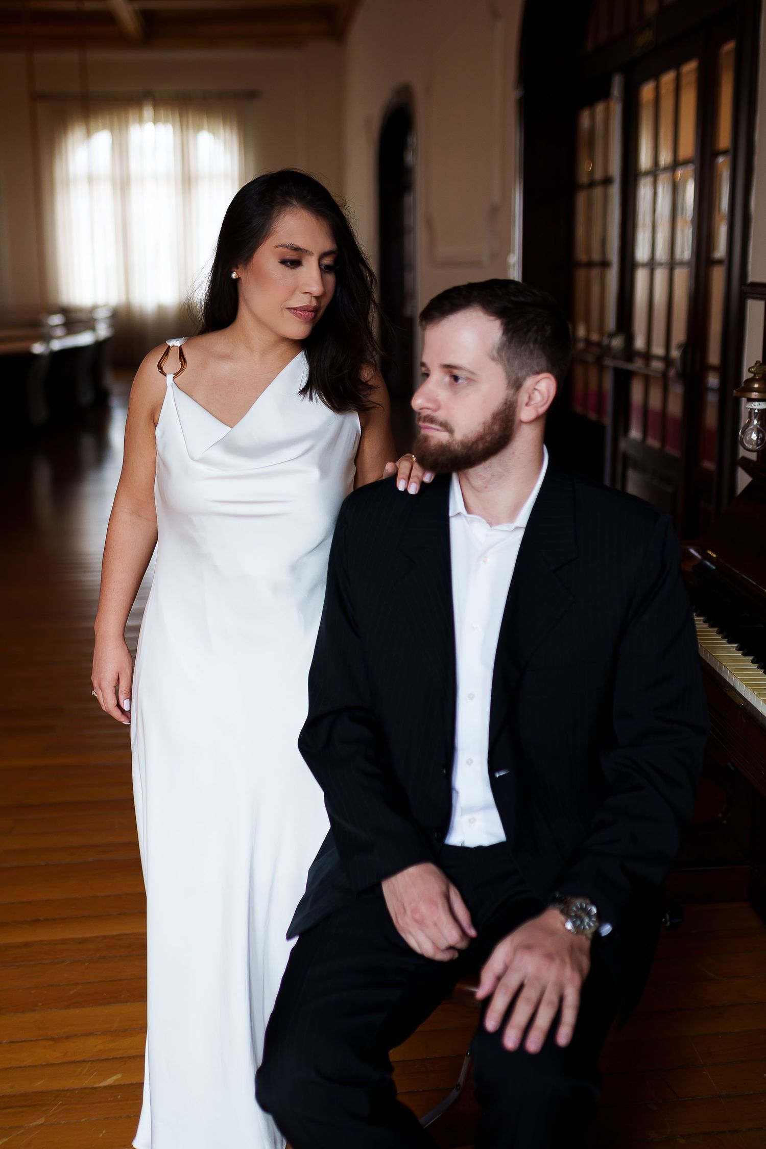 Bride in white gown stands beside groom in black suit kneeling, posing elegantly in a warmly lit room.