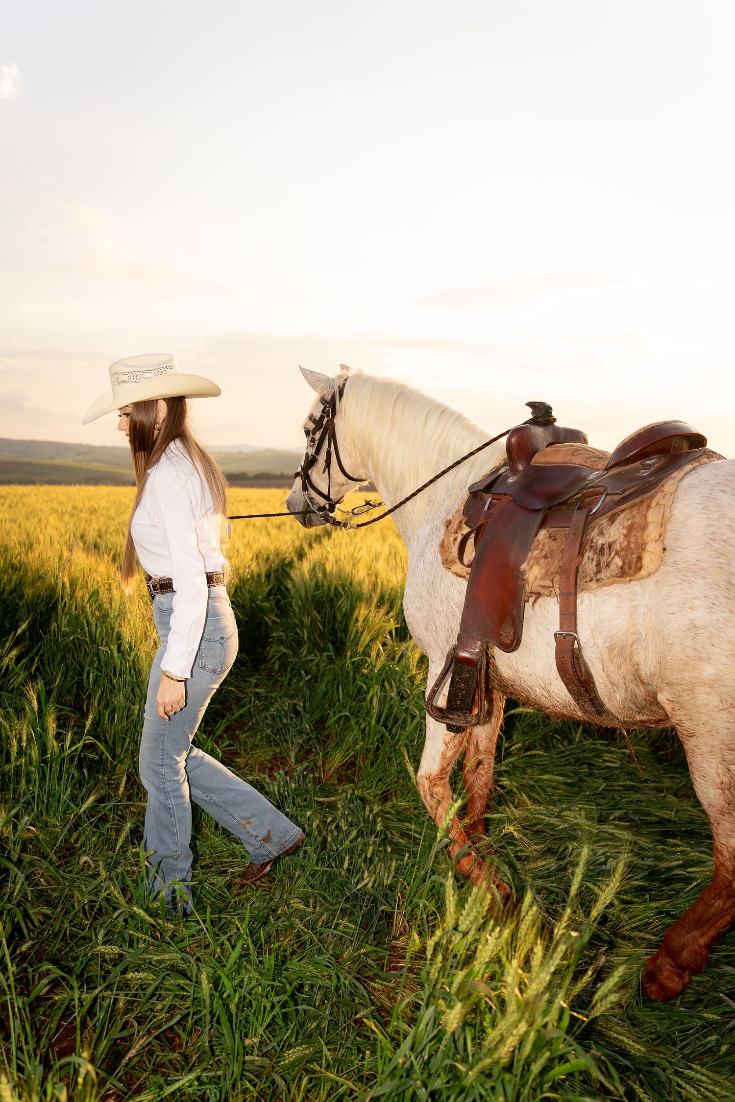 A young woman in a cowboy hat and jeans leads a white saddled horse through a lush green field at golden hour.