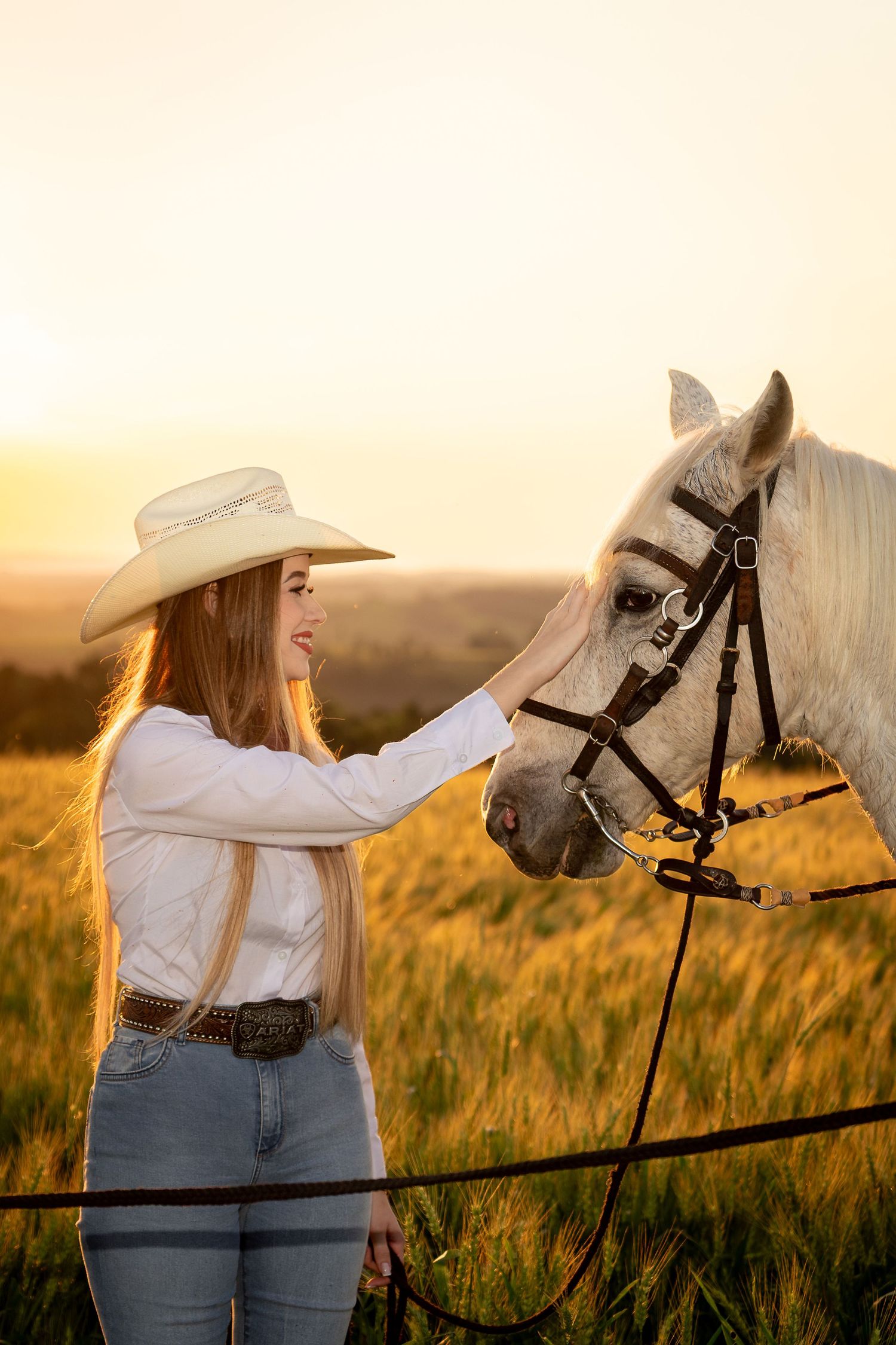 A young woman in a cowboy hat and jeans gently pets a white horse in a golden sunset field.