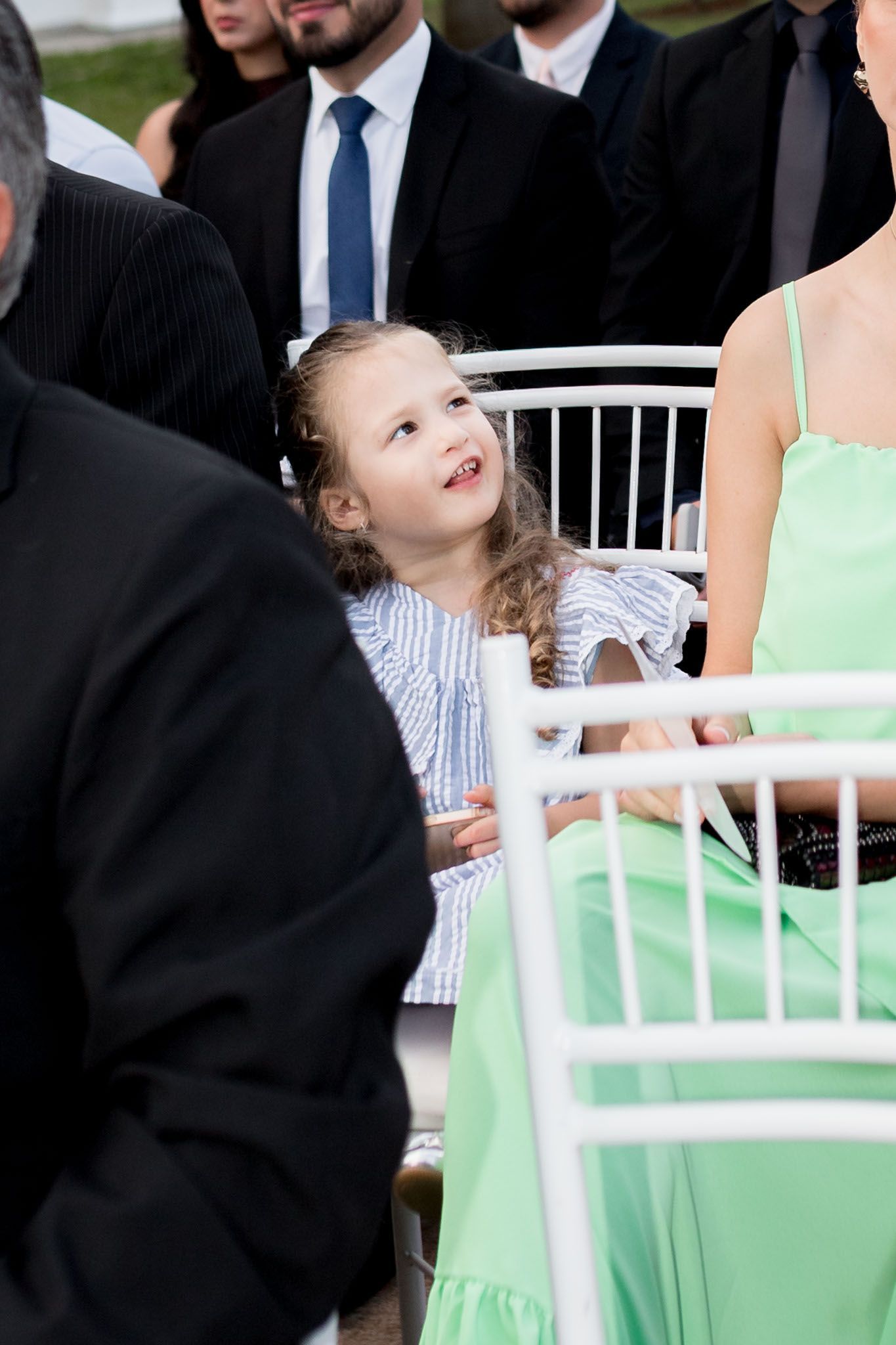 Adorable young girl in striped dress looks up smiling while seated among wedding ceremony guests.