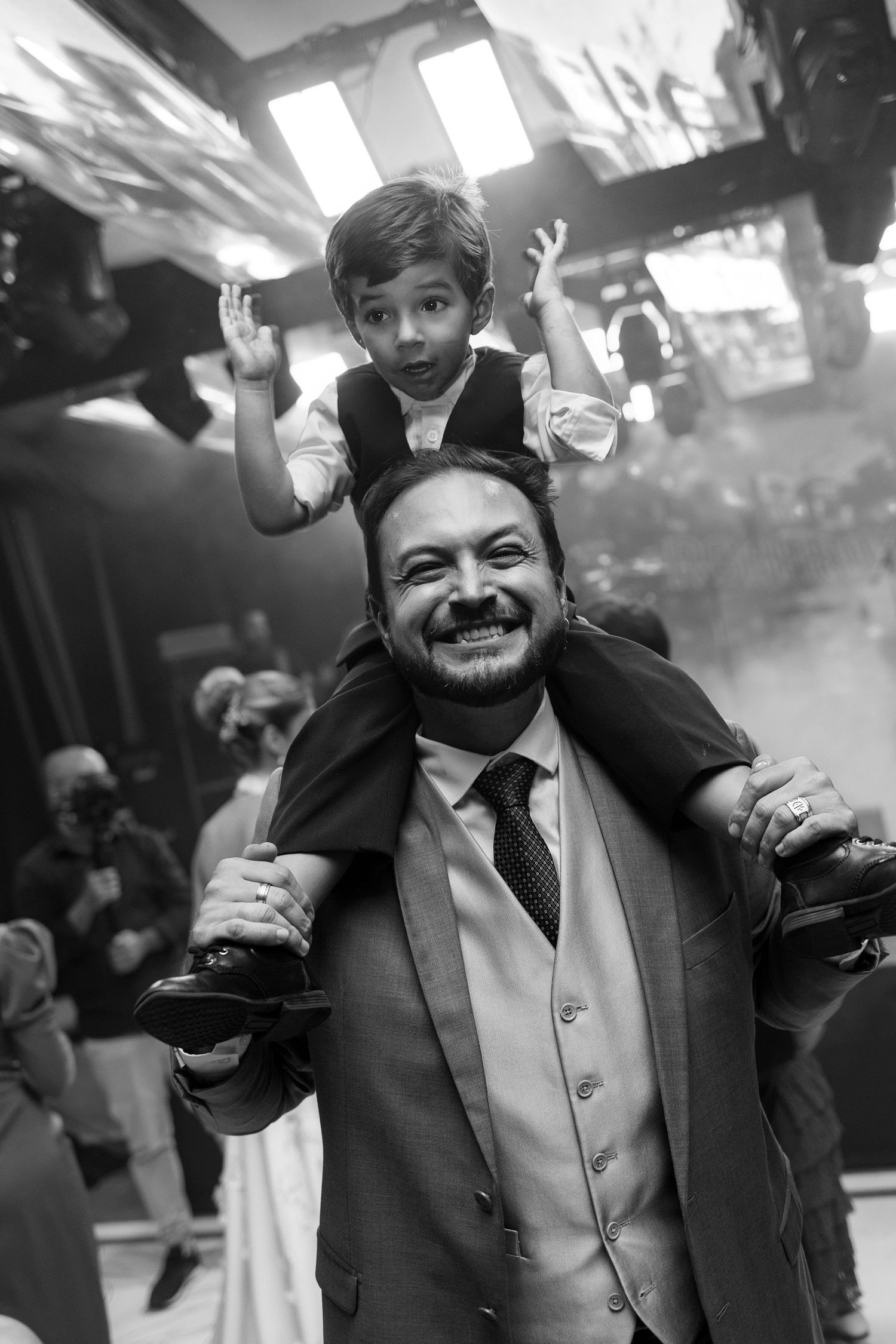 Black and white photo of smiling man carrying excited young boy on his shoulders at a wedding celebration.