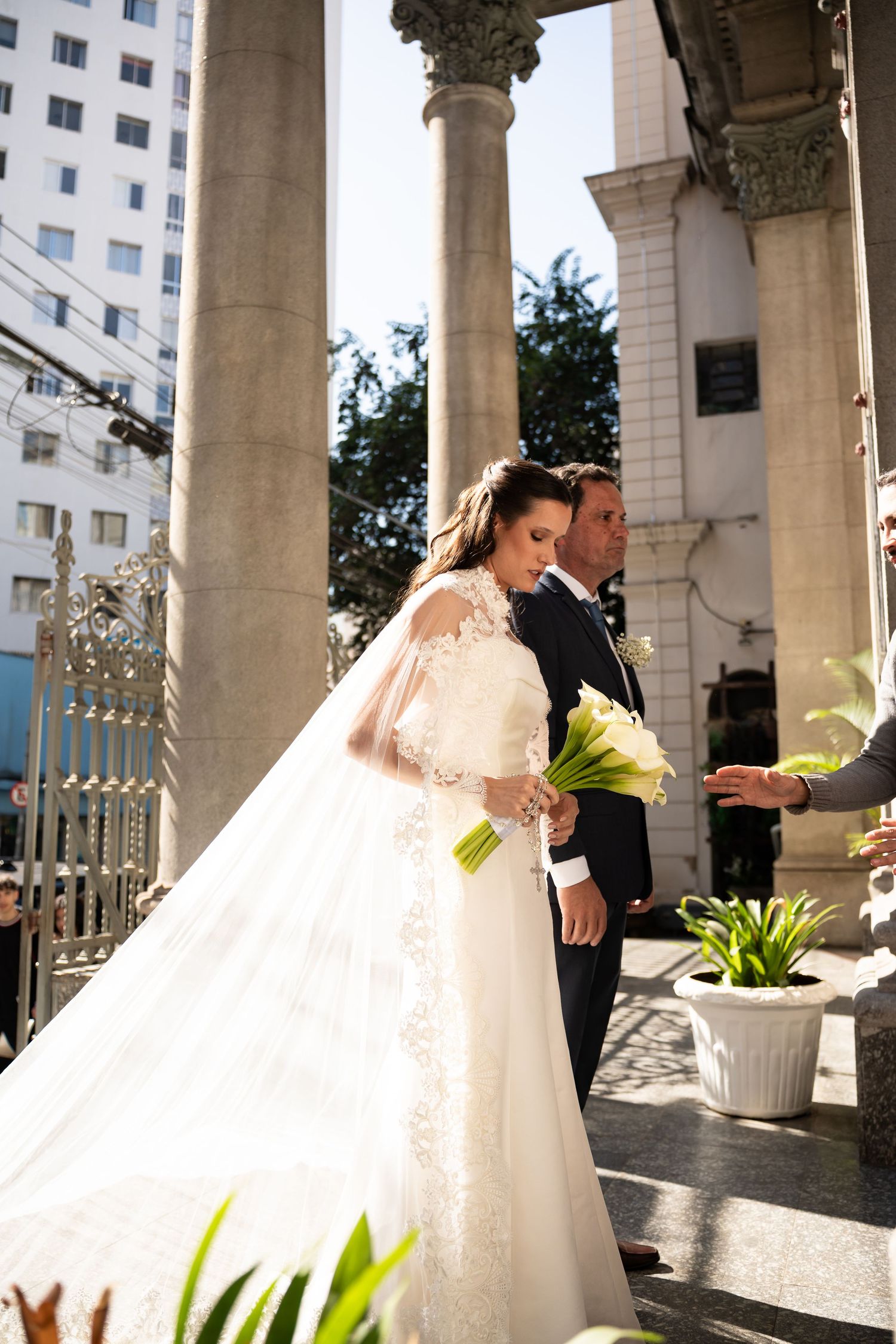 Bride in lace wedding gown holding bouquet stands with groom in black suit beside stone columns at church entrance.