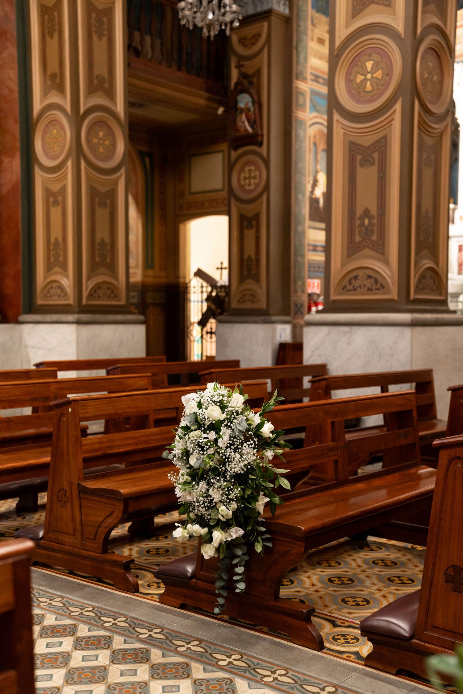 Ornate church interior with wooden pews adorned with white floral arrangement, featuring decorative painted ceilings and marble columns.