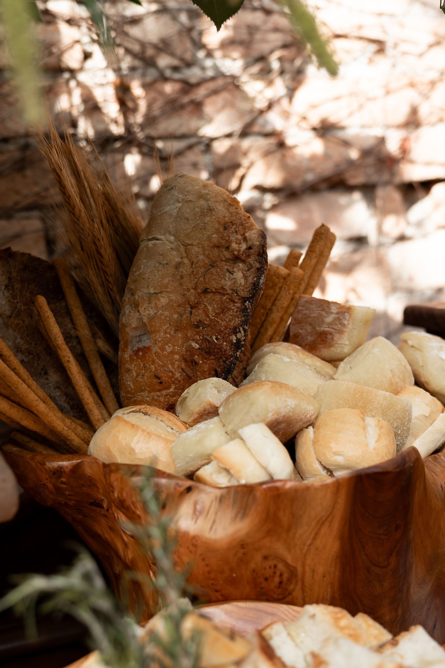 Rustic wooden bowl filled with small bread rolls and cinnamon sticks, displayed outdoors with stone background.