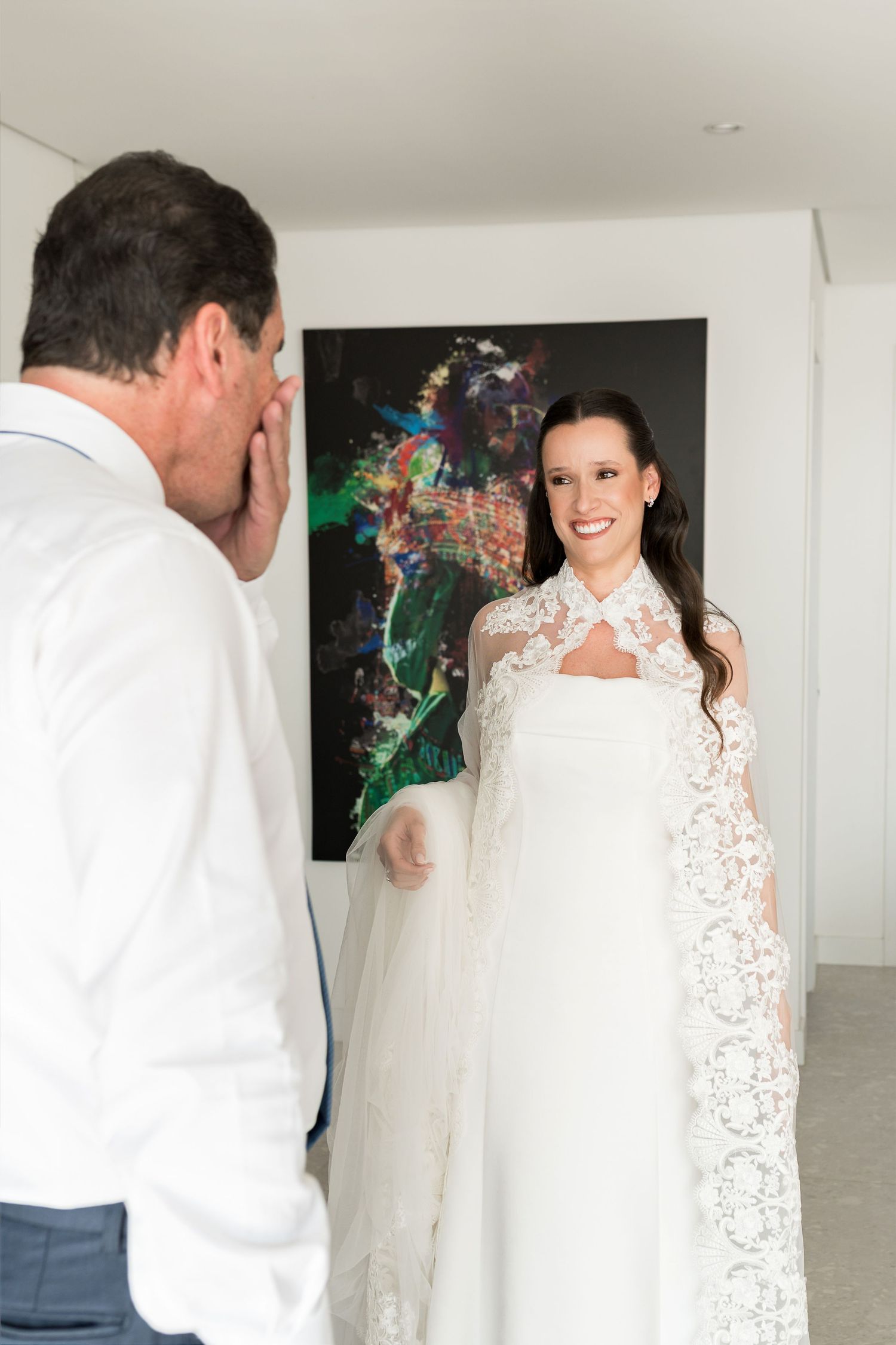 Bride in lace wedding gown and cape smiles at emotional man in white shirt during a heartfelt first look moment.