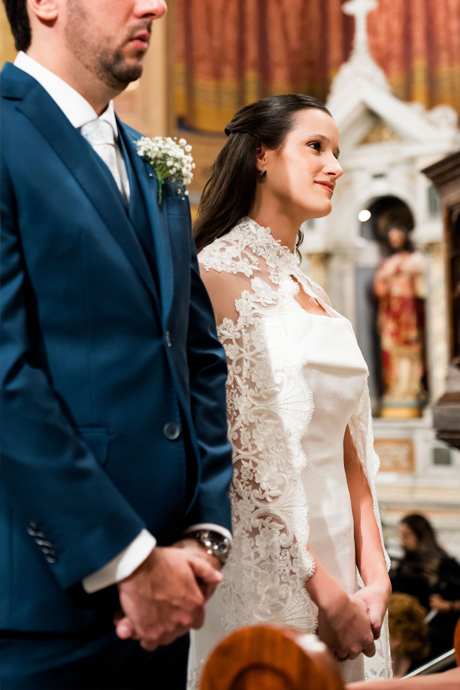 Bride in lace wedding dress holding hands with groom in blue suit during church ceremony.