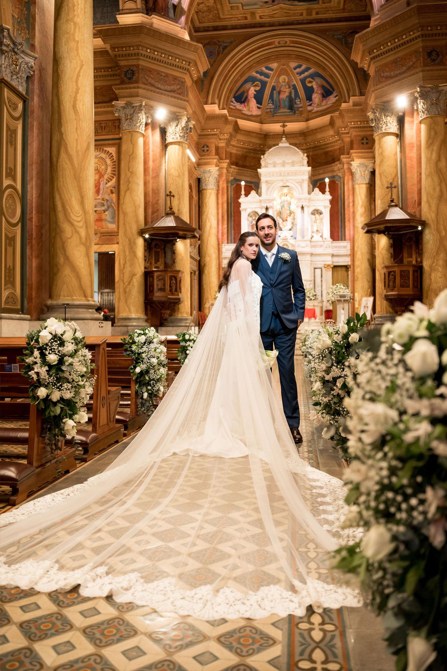Bride and groom standing in an ornate church aisle, bride wearing a long lace cathedral veil, surrounded by white floral arrangements.