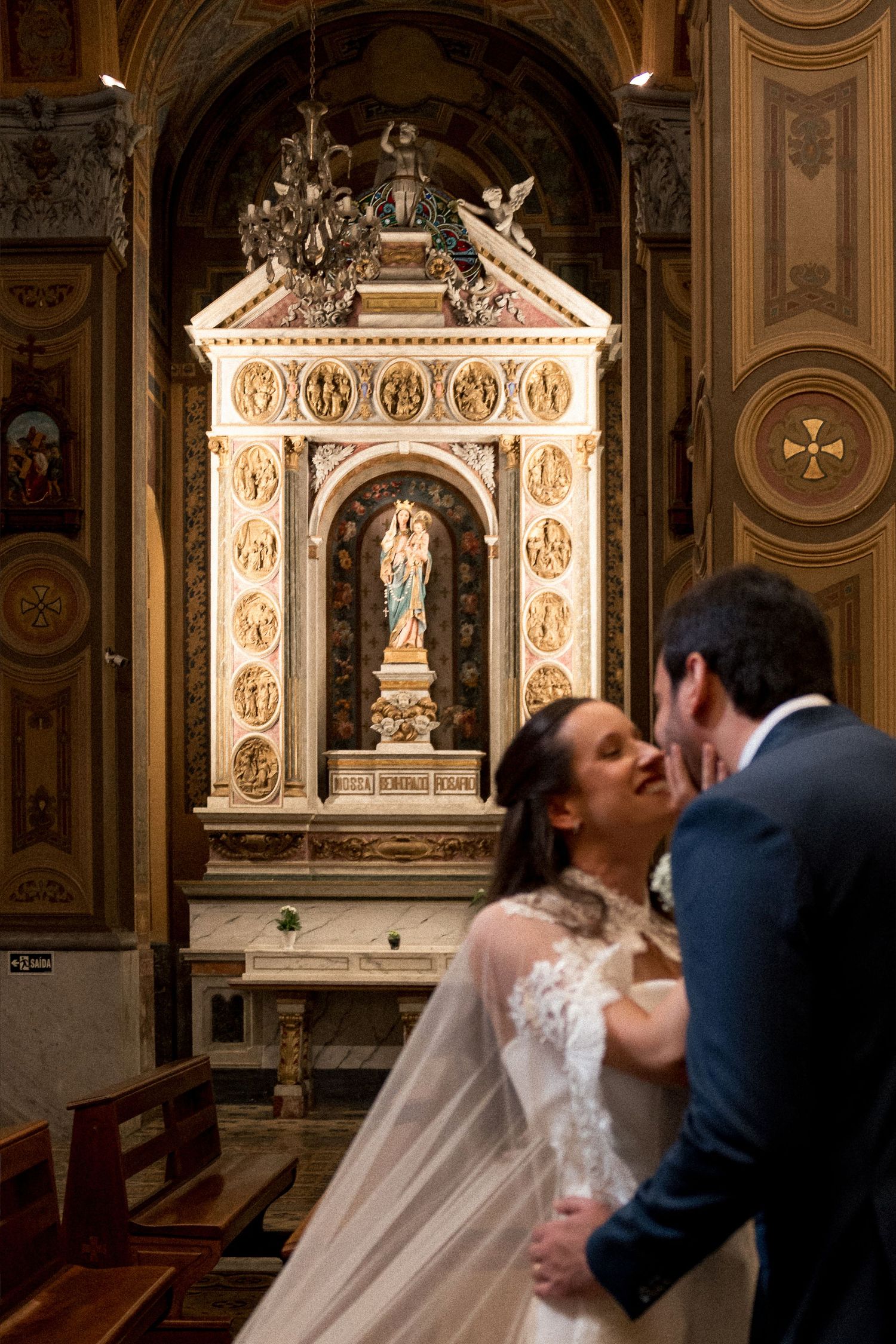 Bride and groom share a kiss in an ornate church with a golden altar and religious statue in the background.