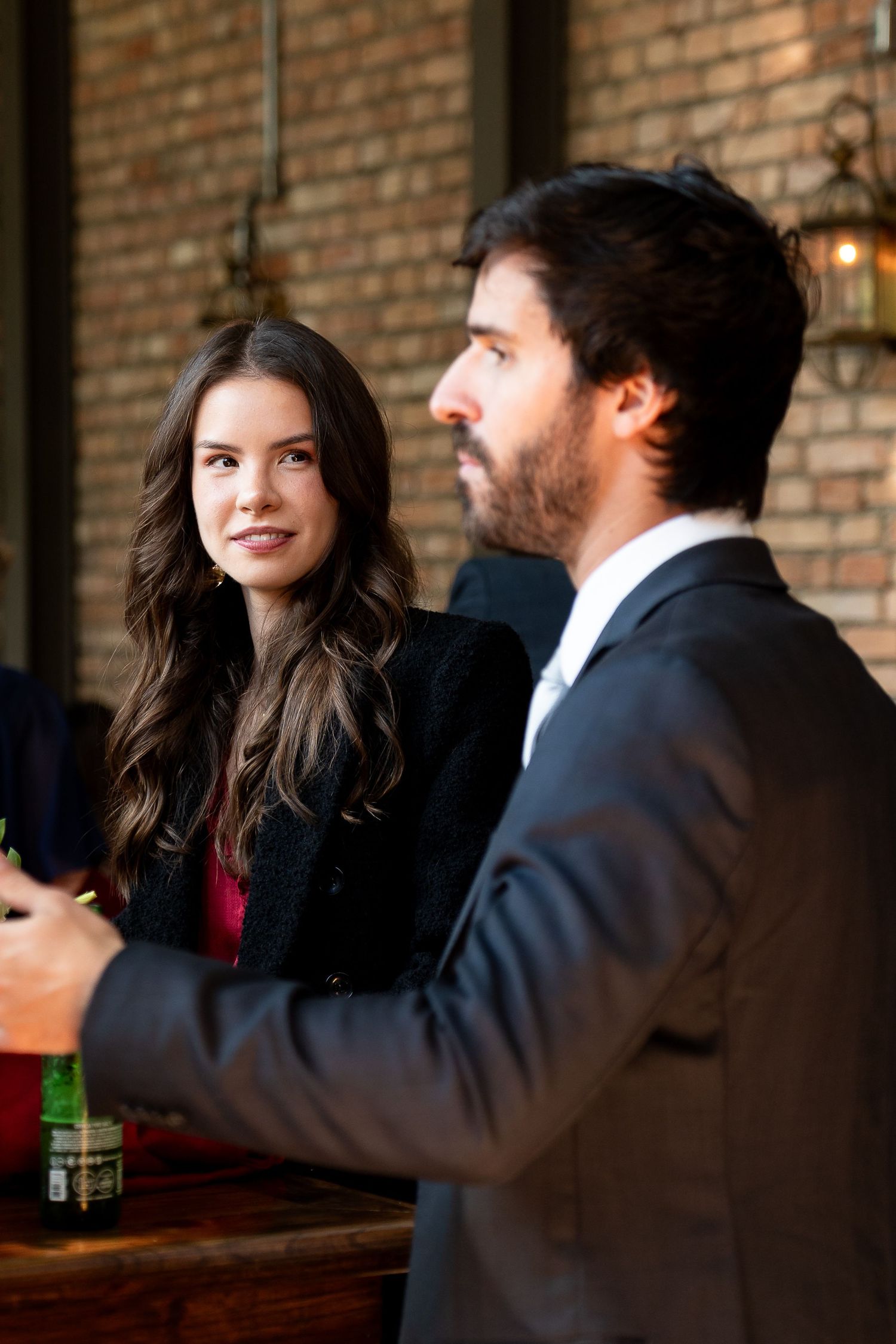 A young woman with long brown hair smiles at a bearded man in a suit at a rustic bar.