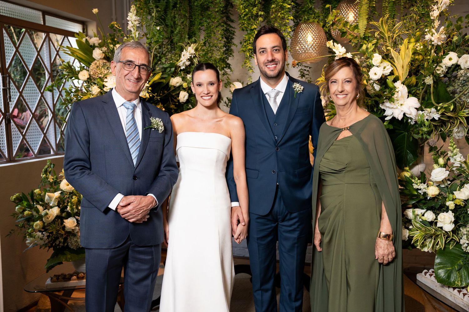 Wedding couple poses with family members amid lush floral arrangements at elegant indoor ceremony venue.