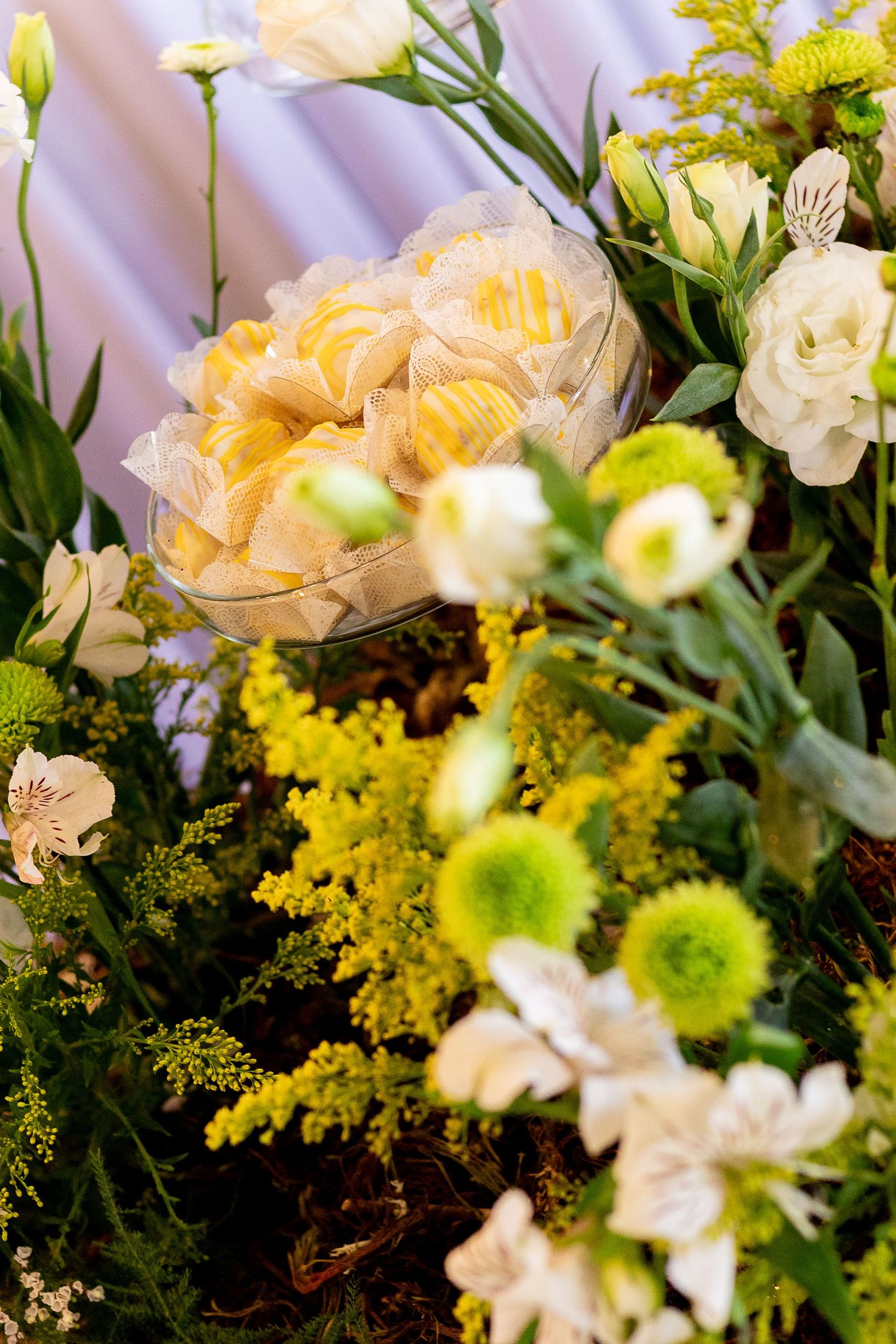 Glass bowl of wrapped yellow candies surrounded by white and green flowers including chrysanthemums and alstroemeria.