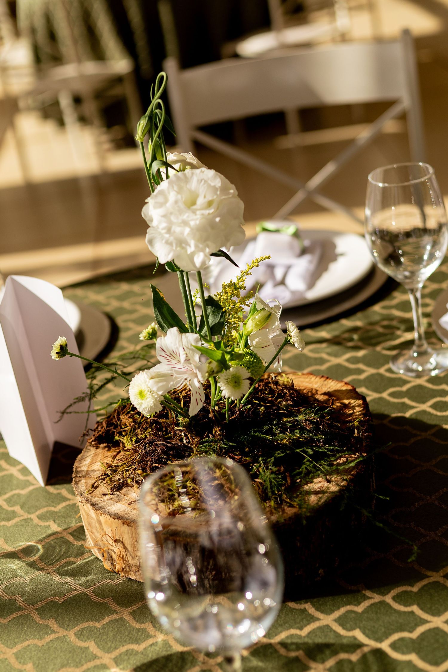Rustic wedding centerpiece with white flowers on wood slice, moss, and wine glasses on green patterned tablecloth.
