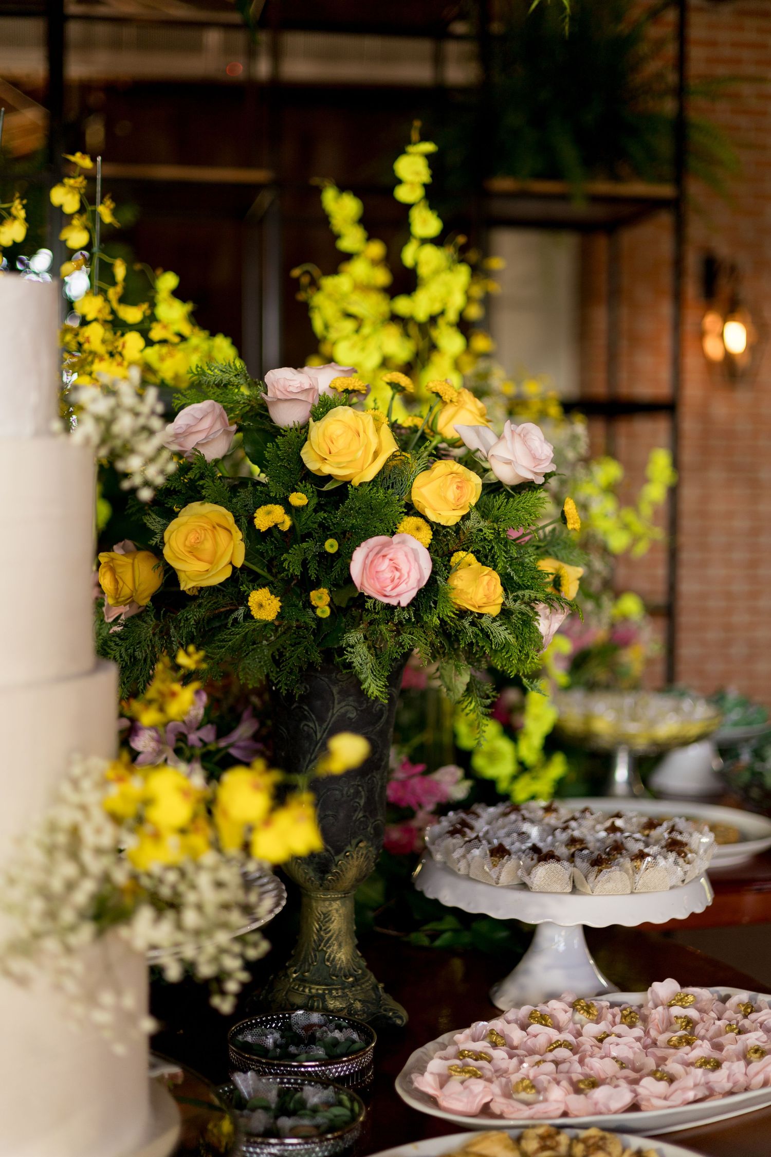 Elegant wedding dessert table with yellow and pink roses, white tiered cake, and assorted sweets on white cake stands.