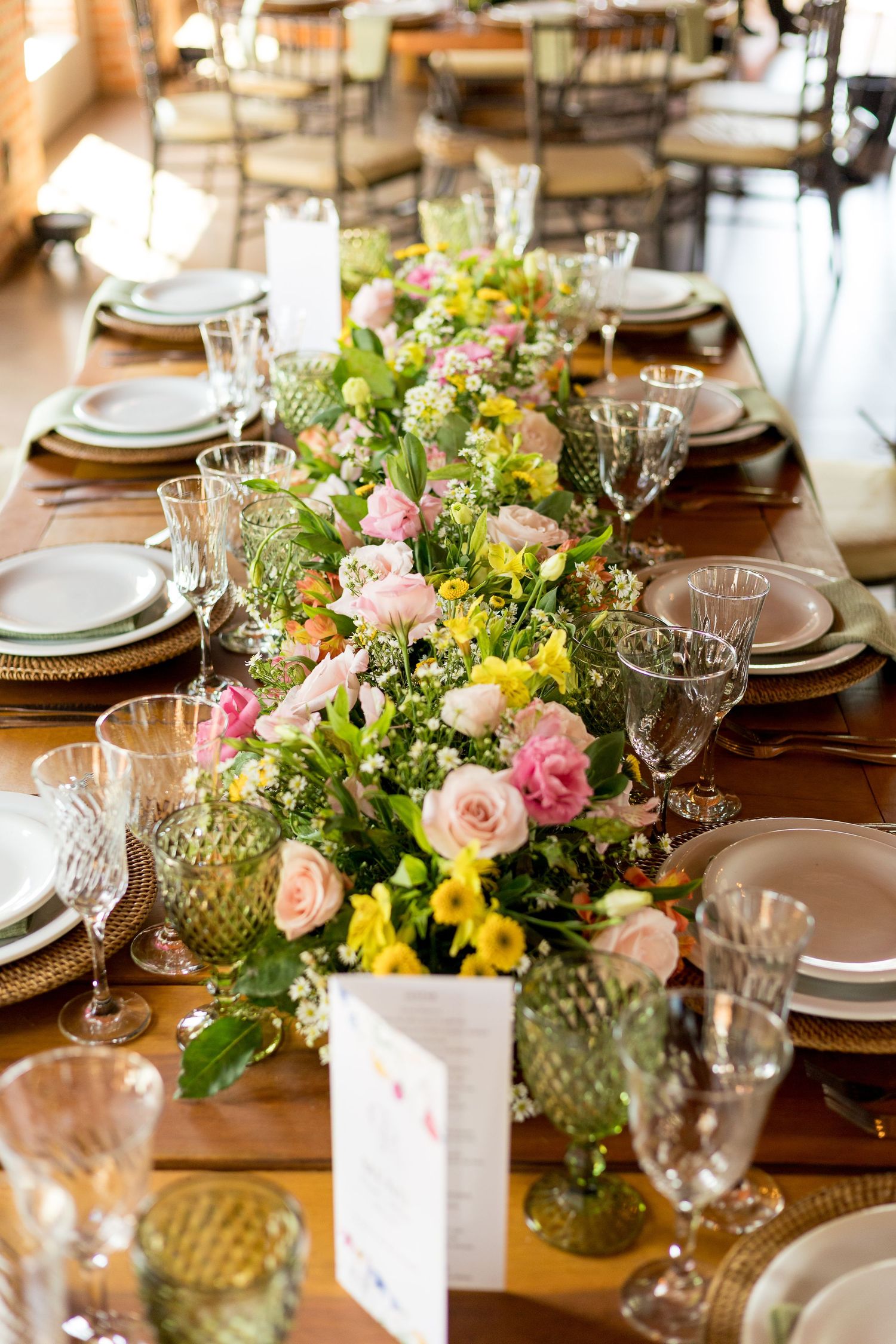 Elegant wedding reception table with lush floral runner of pink roses and yellow blooms, crystal glasses, and white plates on wood table.
