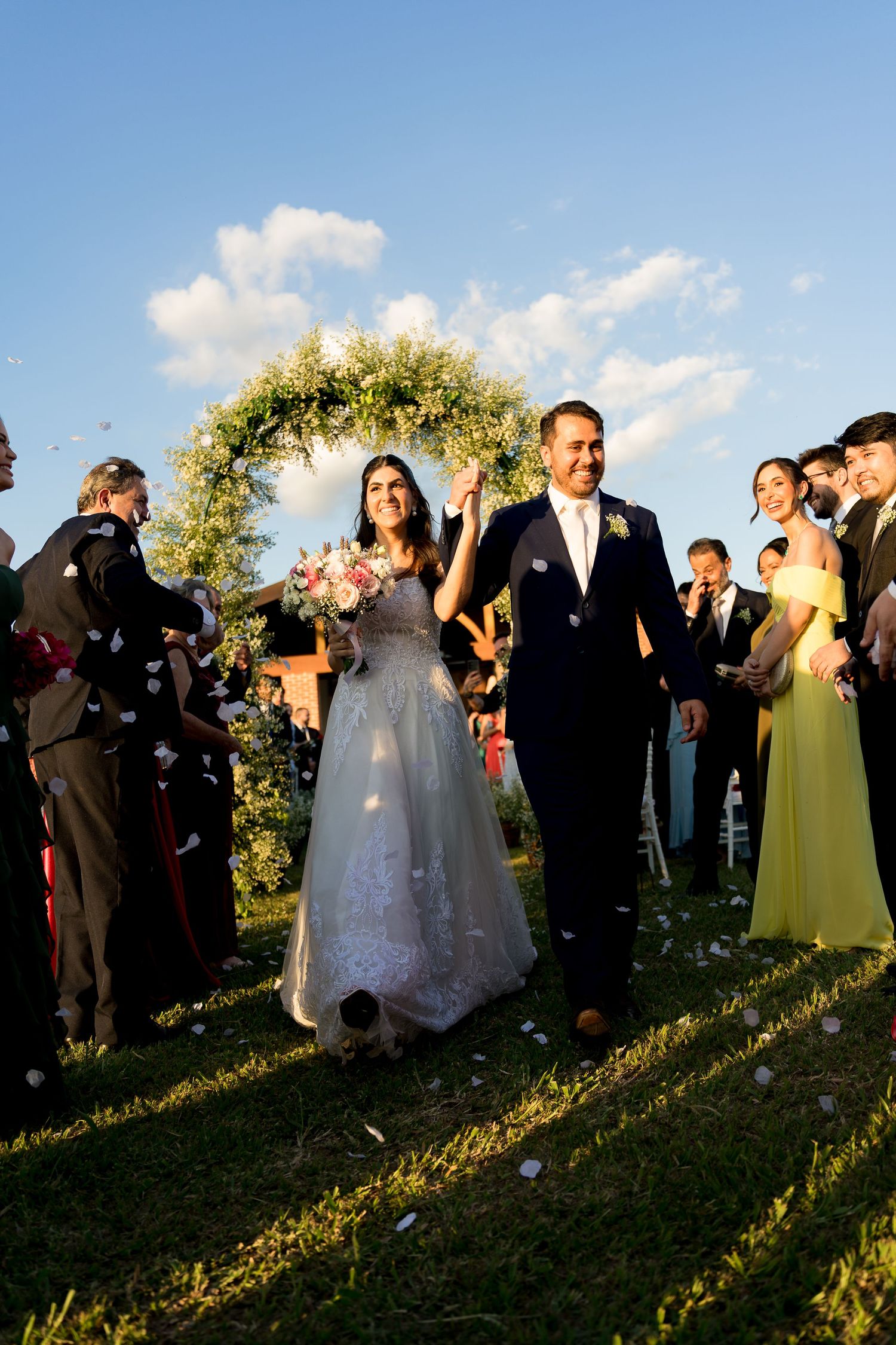 Joyful newlyweds walk down the aisle under a floral arch, greeted by confetti and cheering guests outdoors.
