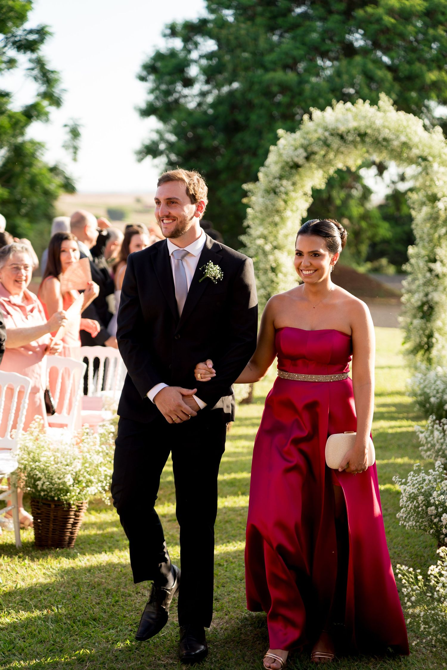 A man in a tuxedo and woman in a red strapless gown walk down a floral wedding aisle outdoors.