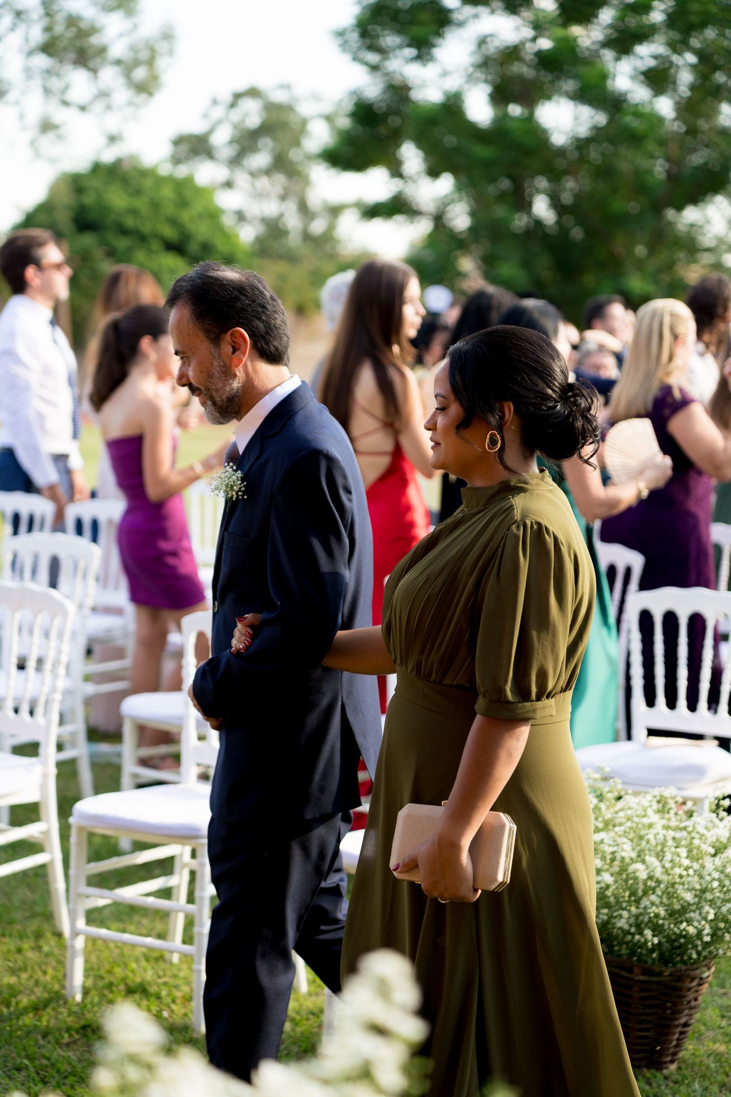 Couple walking arm in arm at an outdoor wedding ceremony with white chairs and guests in colorful attire.