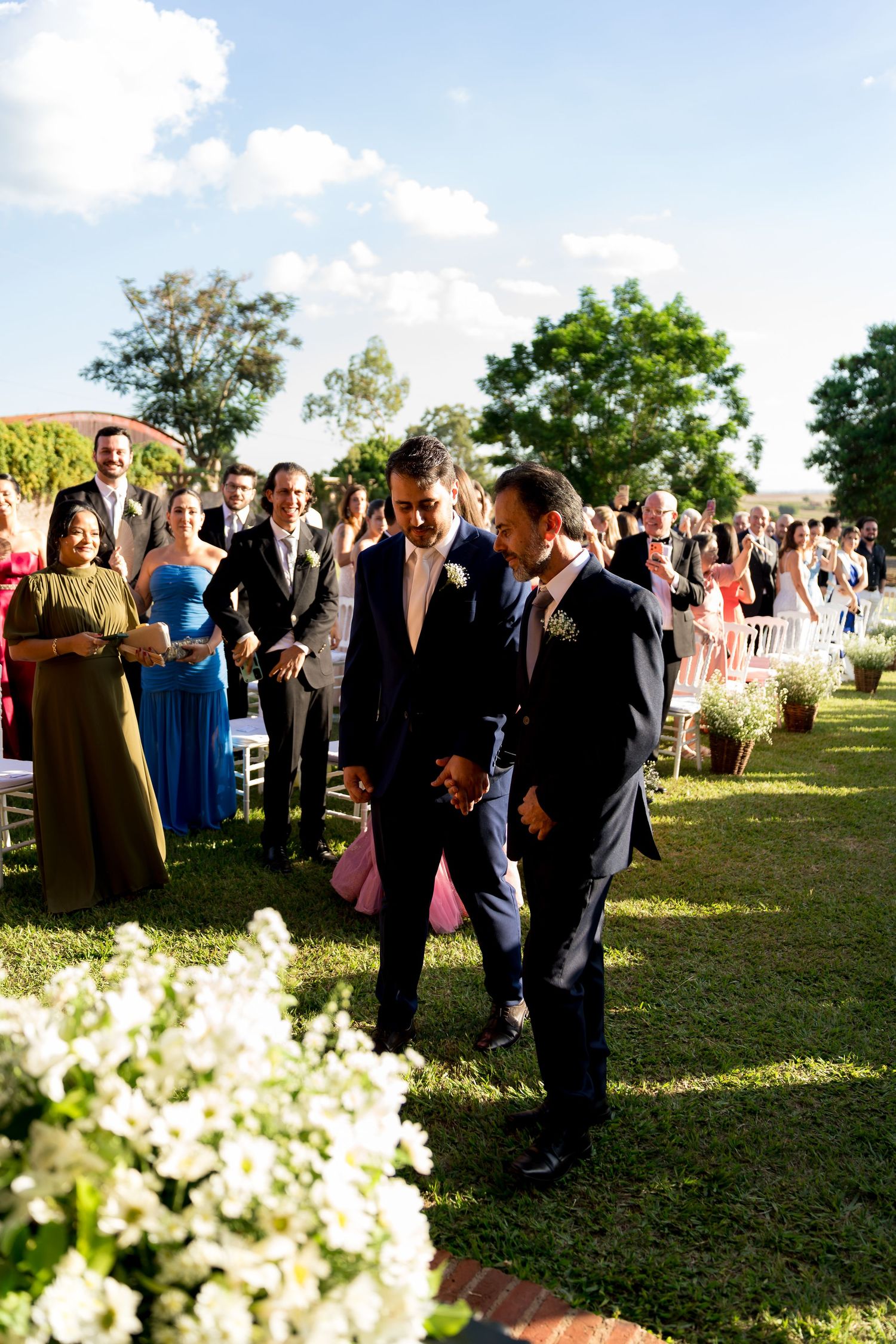 Two grooms walk down the aisle at an outdoor wedding ceremony surrounded by guests and white floral arrangements.