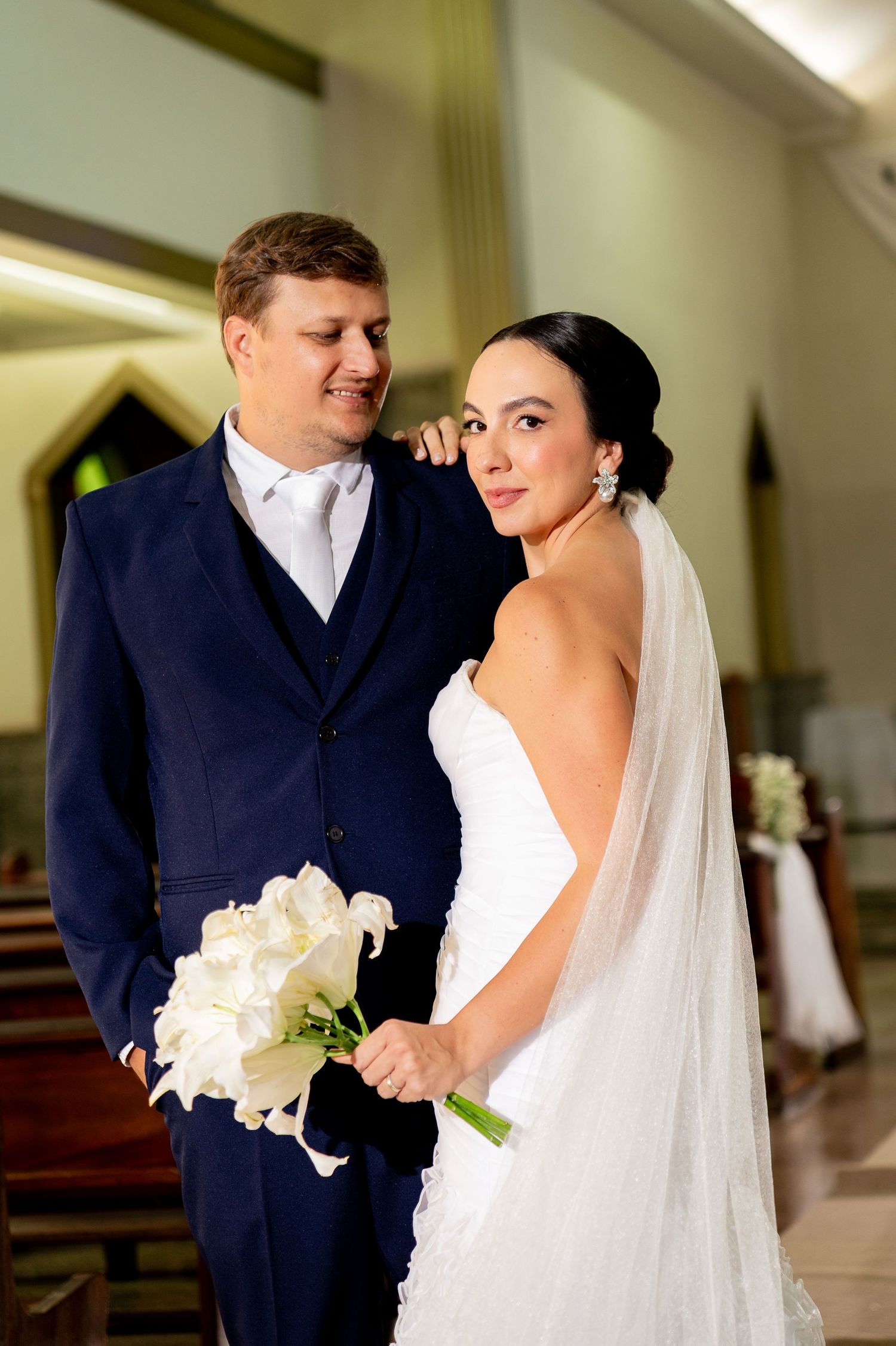 A bride in a white gown and veil holds white flowers beside a groom in a navy suit inside a church.