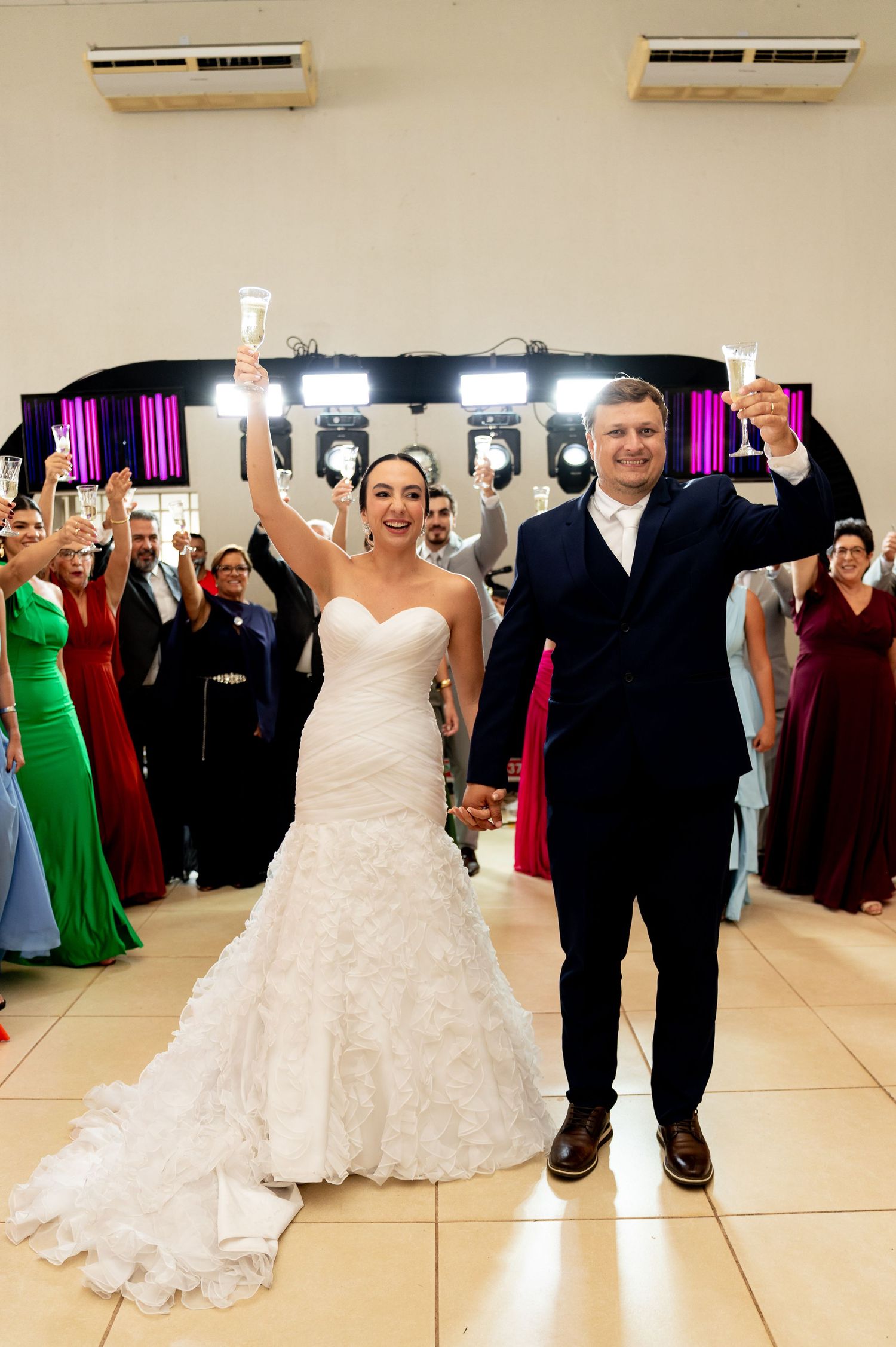 Bride and groom raise champagne glasses at wedding reception, surrounded by guests in formal attire on dance floor.