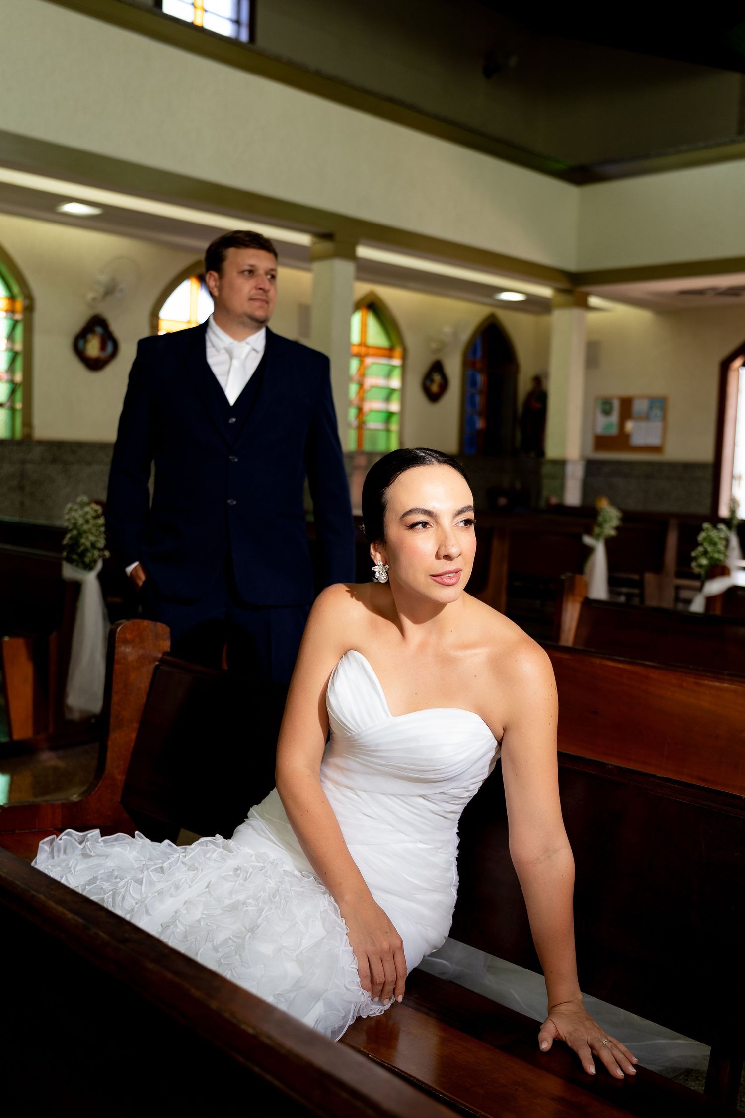 Bride in white strapless gown sits on church pew while groom in navy suit stands behind her.
