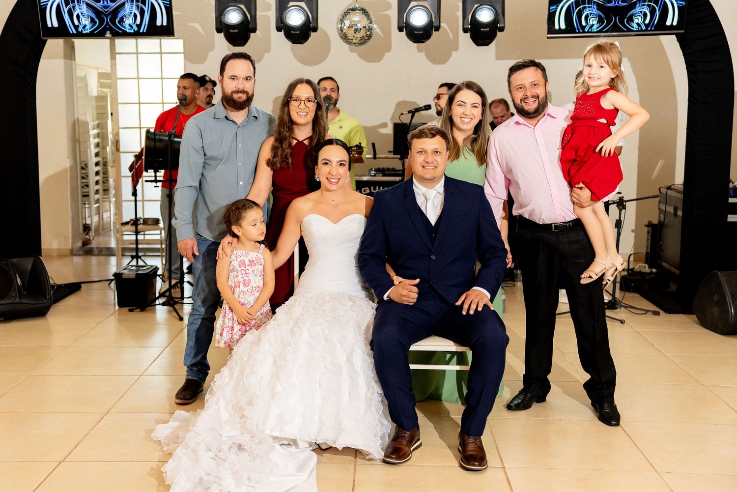 Wedding couple poses with family members in elegant reception venue with disco ball and stage lighting.
