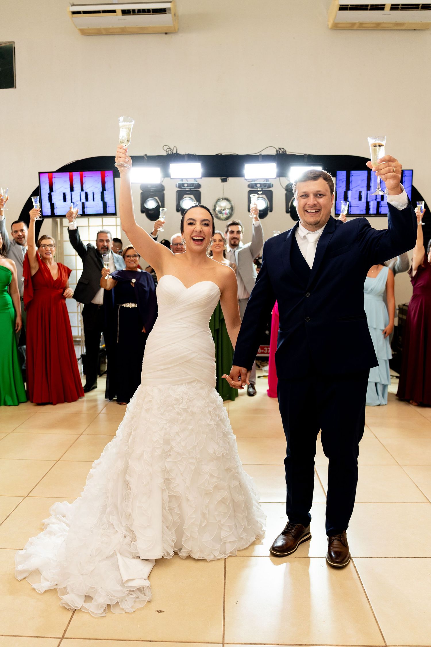 Bride and groom raising champagne glasses during wedding reception toast with guests celebrating in background.