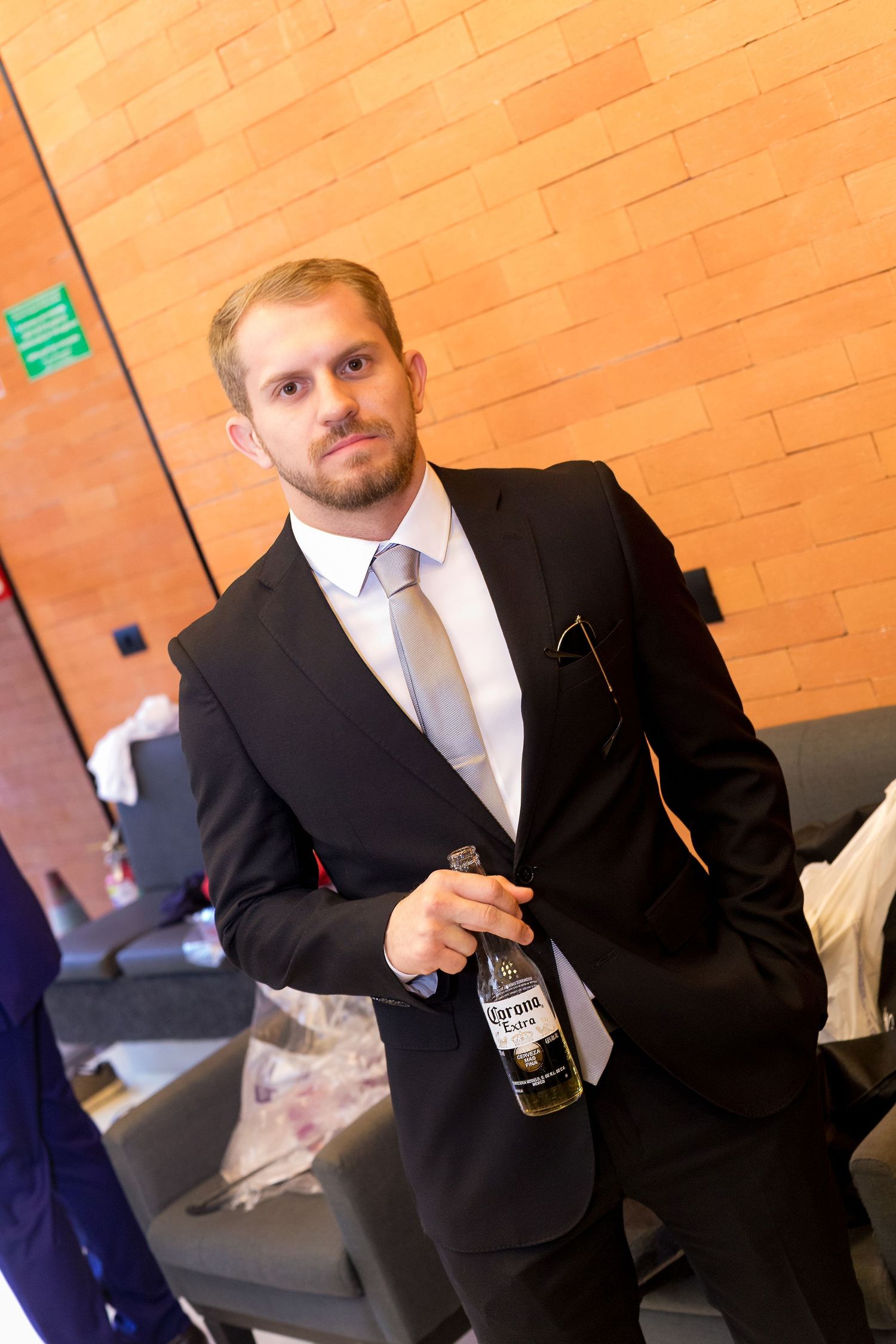 Man in black suit holding Corona beer bottle standing near brick wall at formal event.