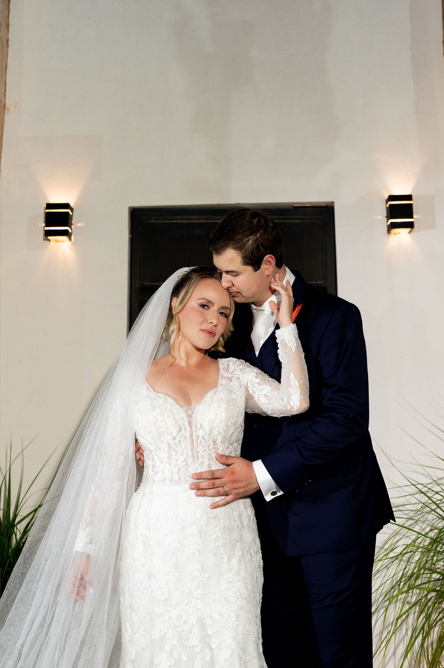 Bride in lace gown with veil and groom in navy suit share an intimate moment by a white wall with sconce lights.