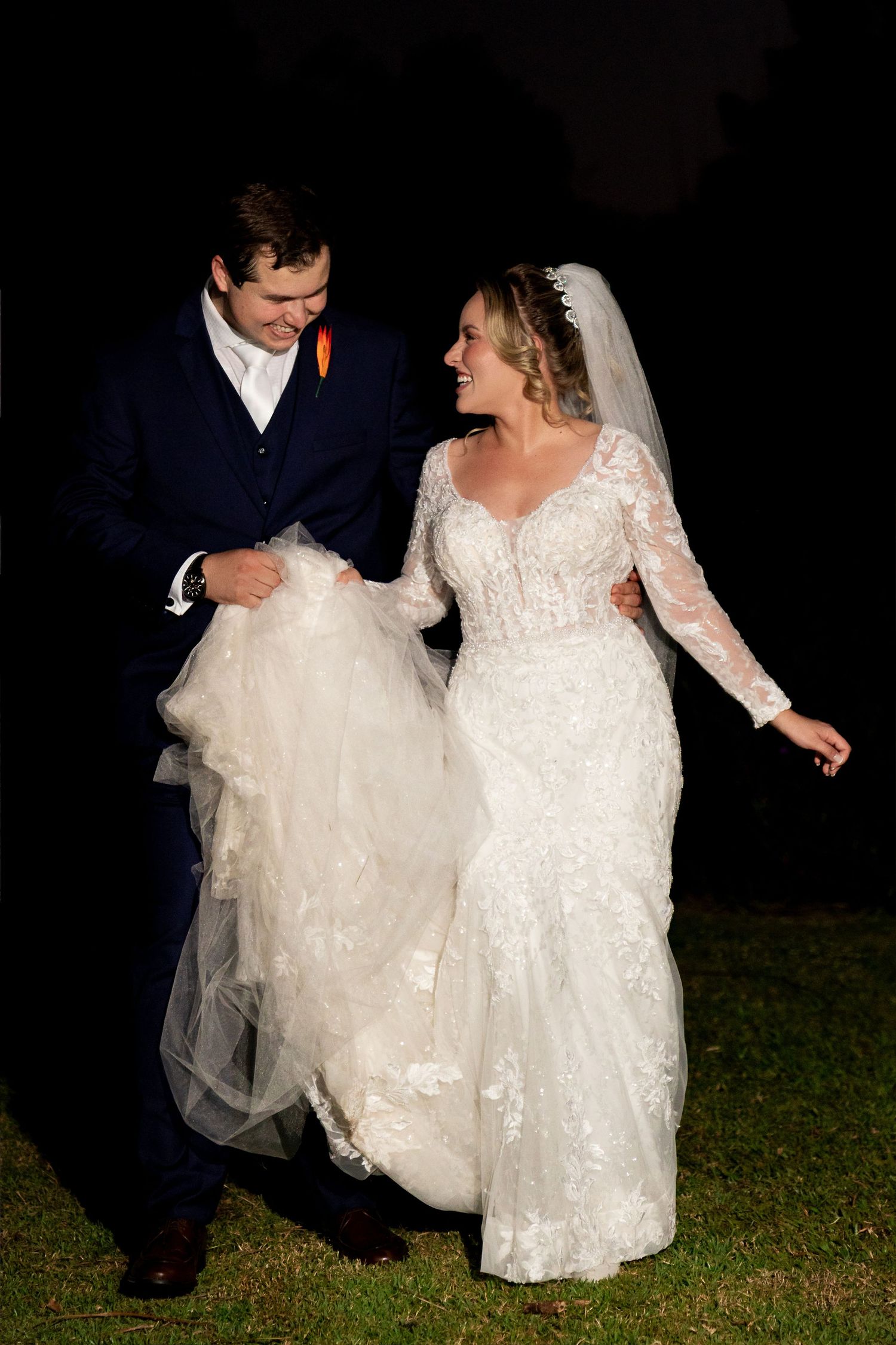 Bride in lace gown and groom in navy suit walk together smiling at night during their wedding celebration.
