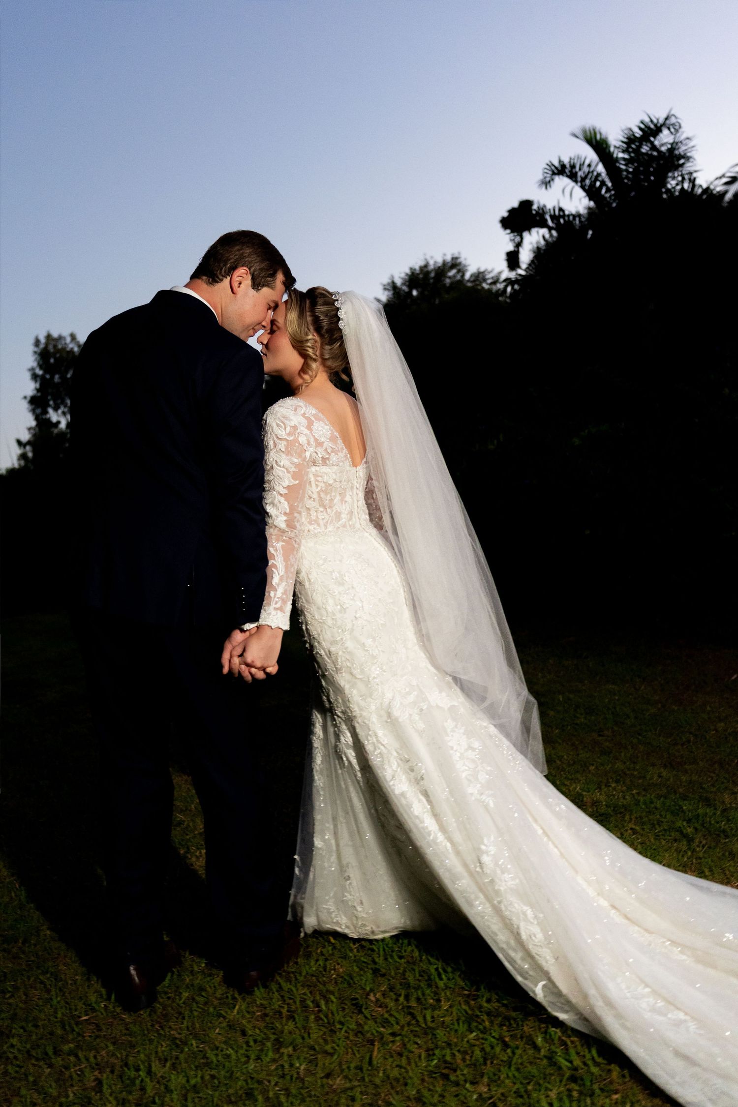 Bride and groom share a romantic forehead touch in golden hour light, her lace gown and veil trailing on green lawn.