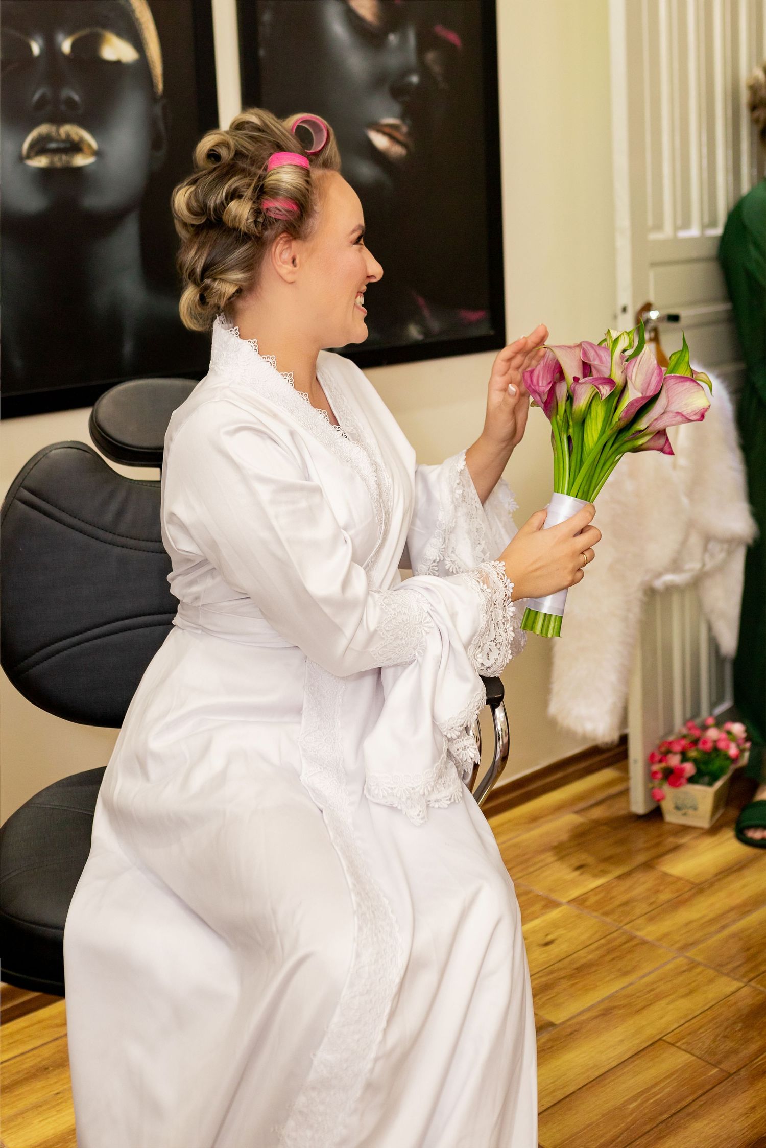 Bride in white robe with pink hair curlers holds a pink tulip bouquet while getting ready at a hair salon.