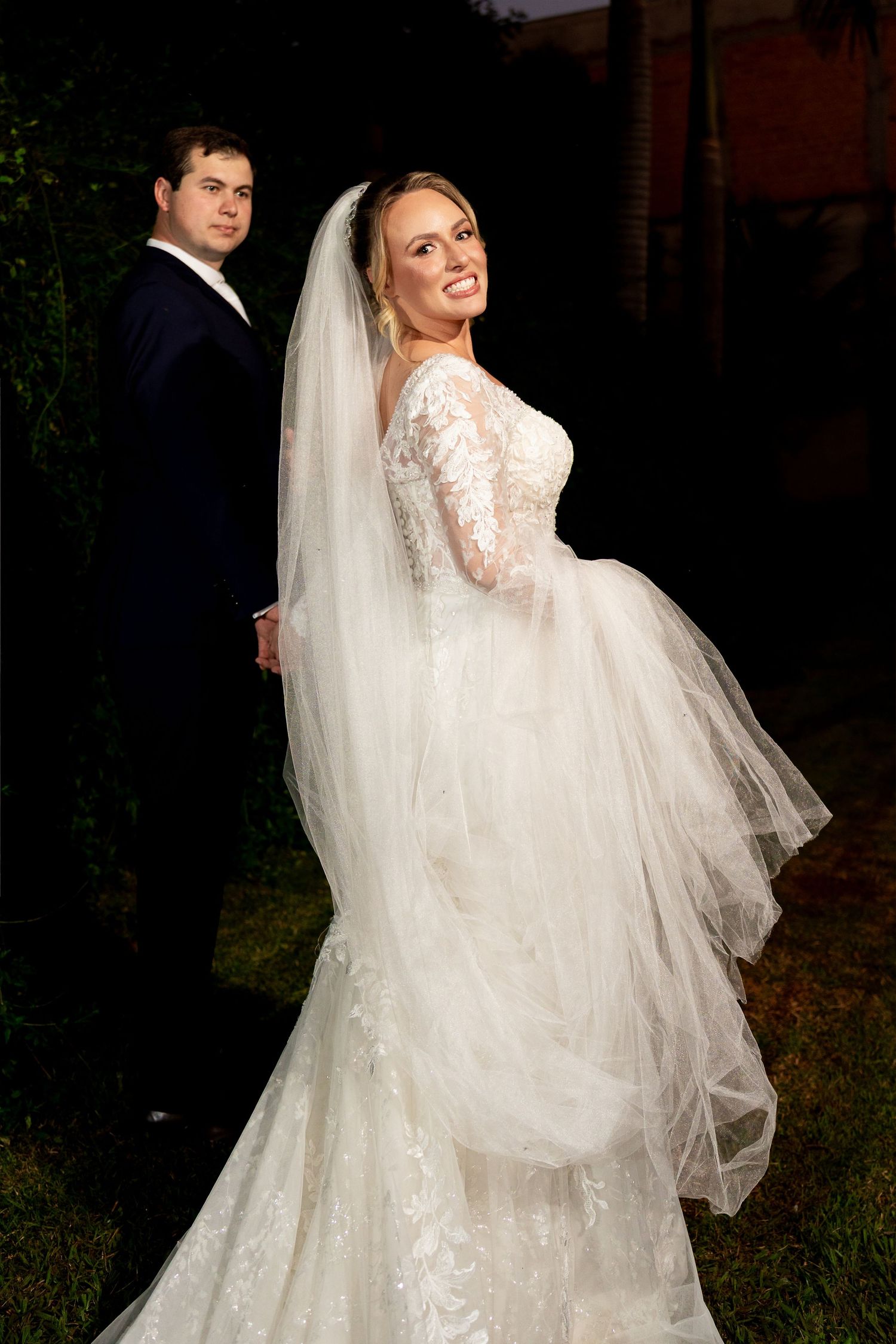 Bride in lace wedding gown and veil holds groom's hand outdoors at night, smiling elegantly at camera.