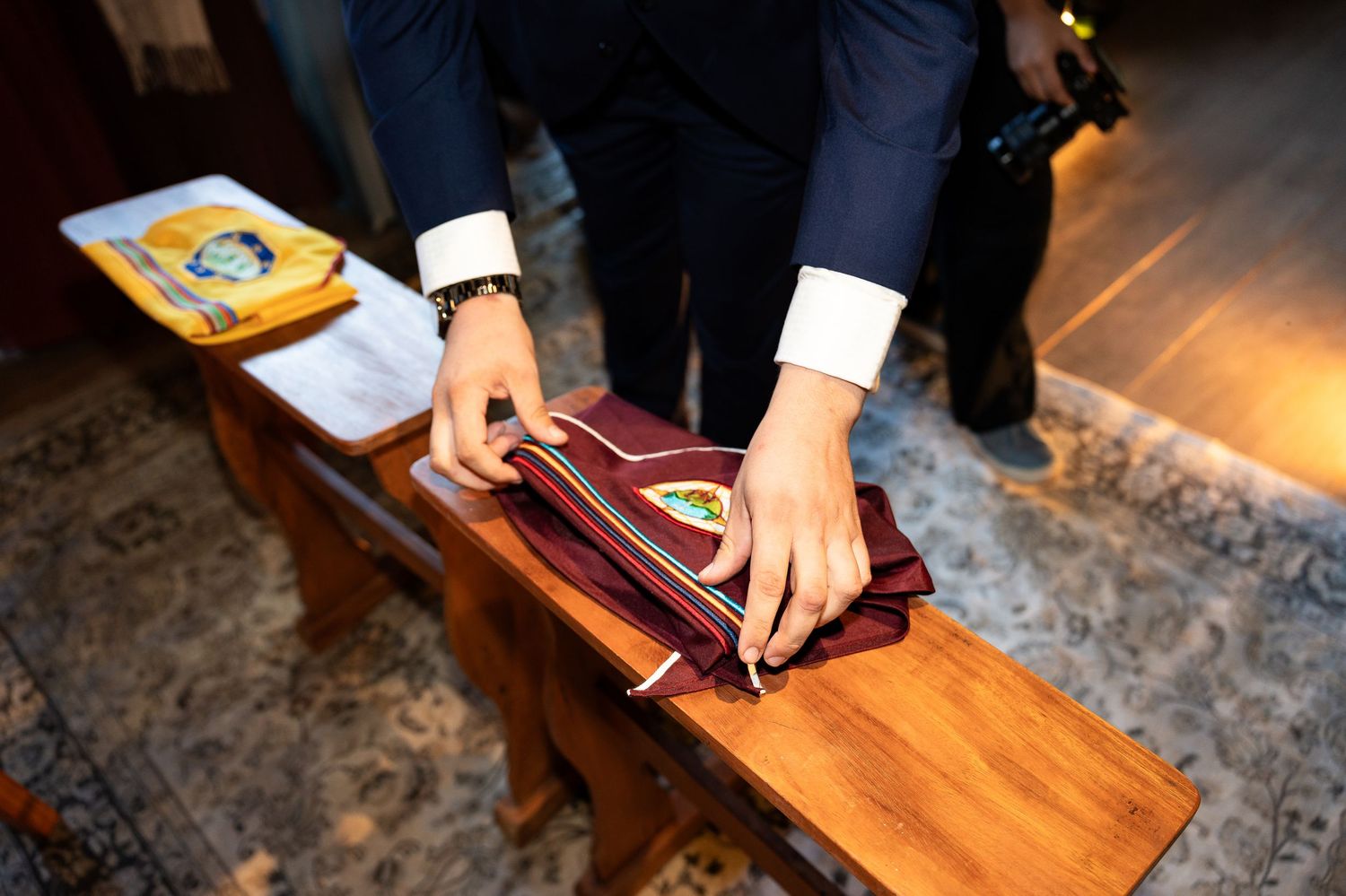 Man in suit carefully folding a burgundy ceremonial sash with emblem on a wooden bench.