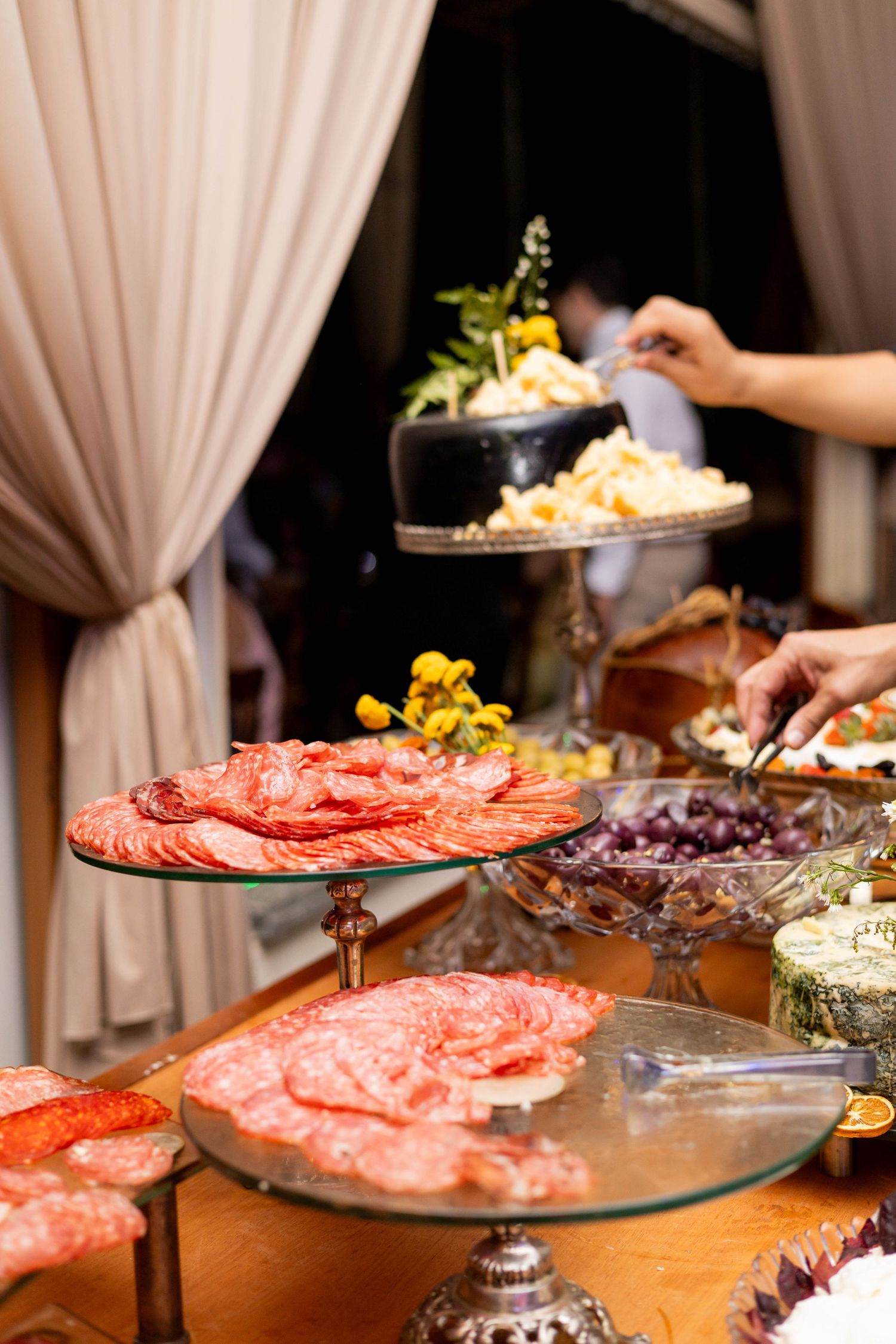 Elegant charcuterie board display with sliced salami, cheese, and garnishes on tiered glass stands at a catered event.