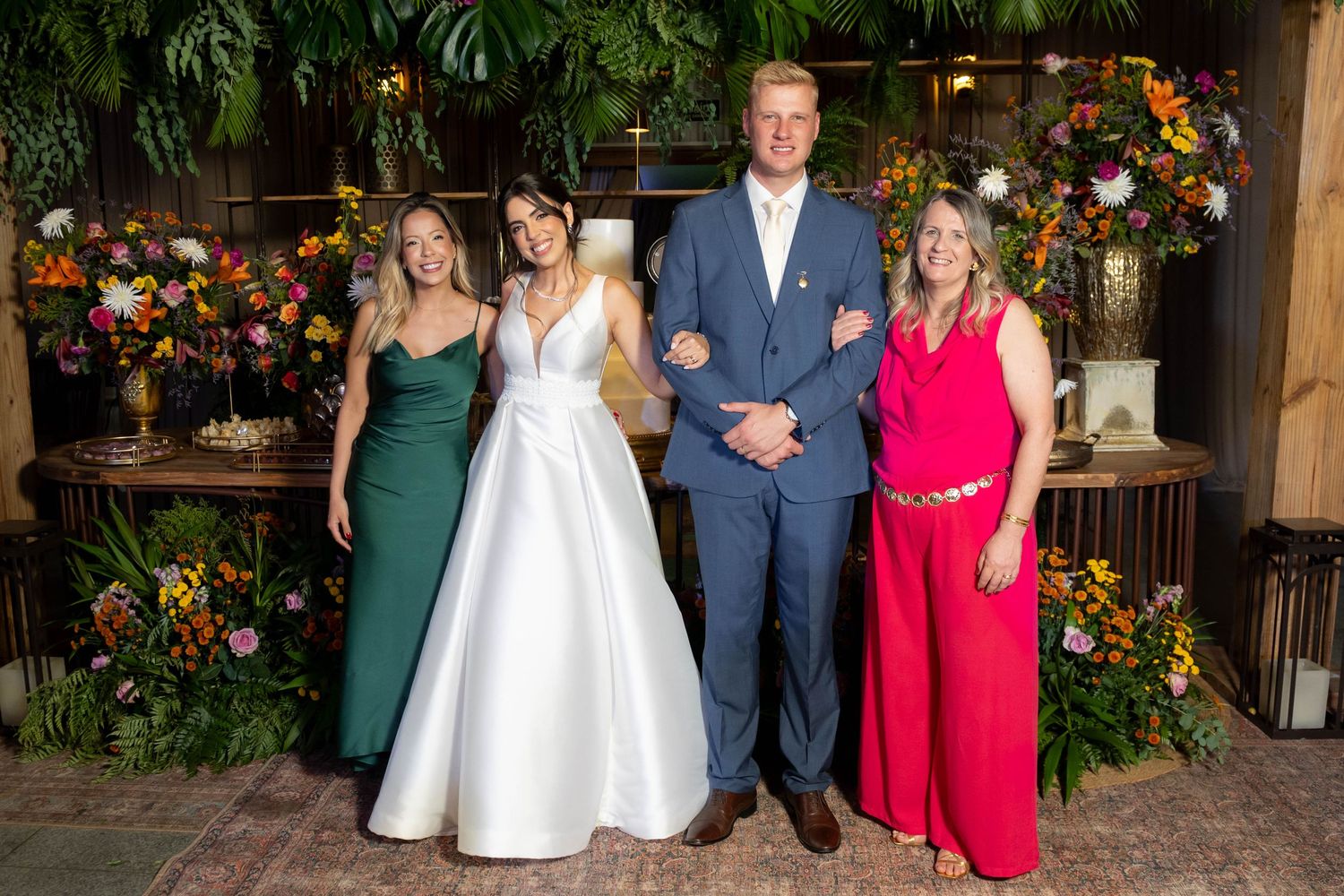 Bride in white gown poses with groom in blue suit and two guests amid colorful floral backdrop at wedding reception.