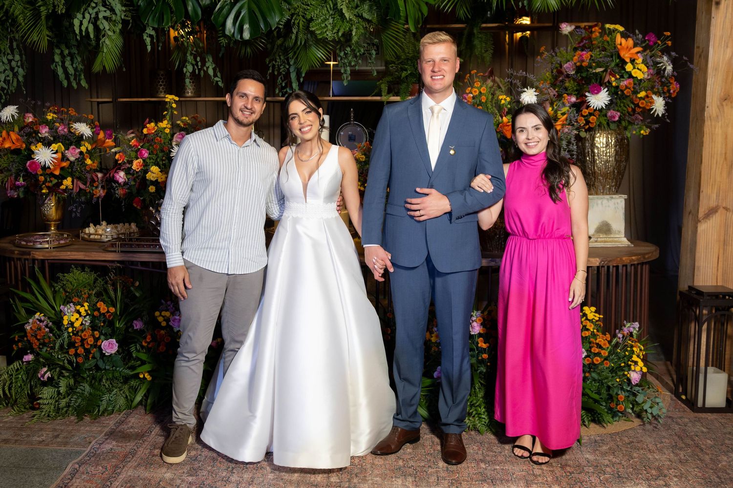 Four people posing together at a wedding celebration, bride in white gown, groom in blue suit, guests in pink and gray.