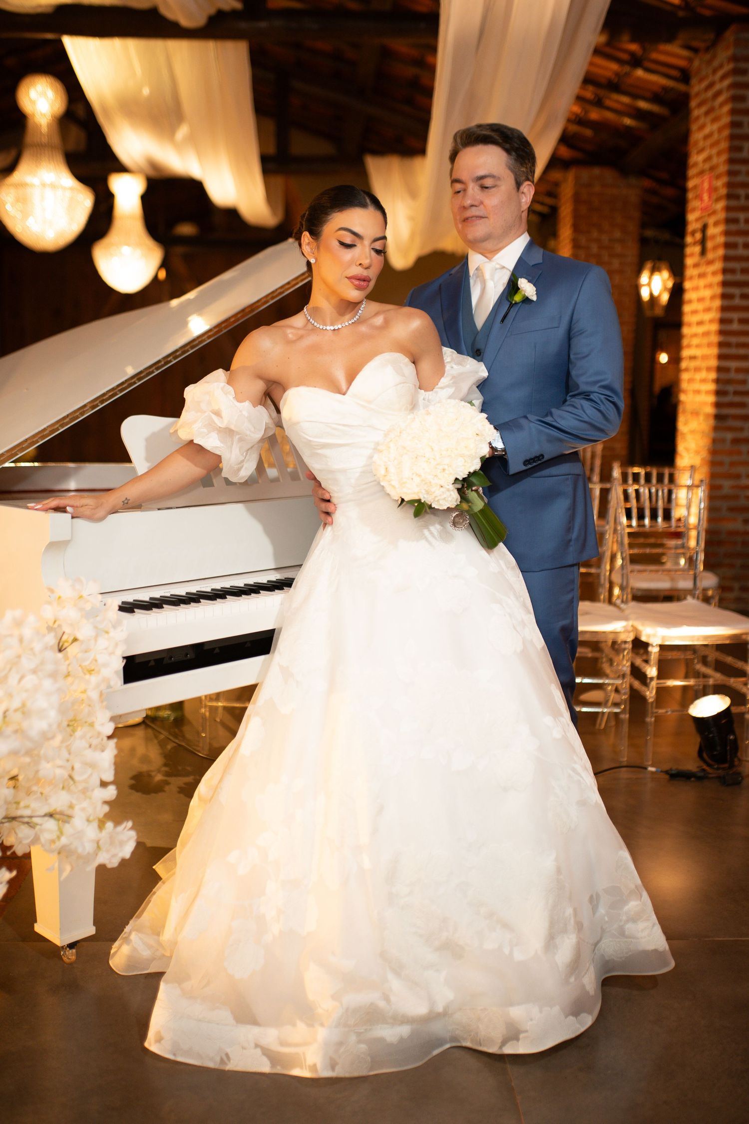 Bride in white ball gown with bouquet stands before white piano, groom in blue suit behind her.
