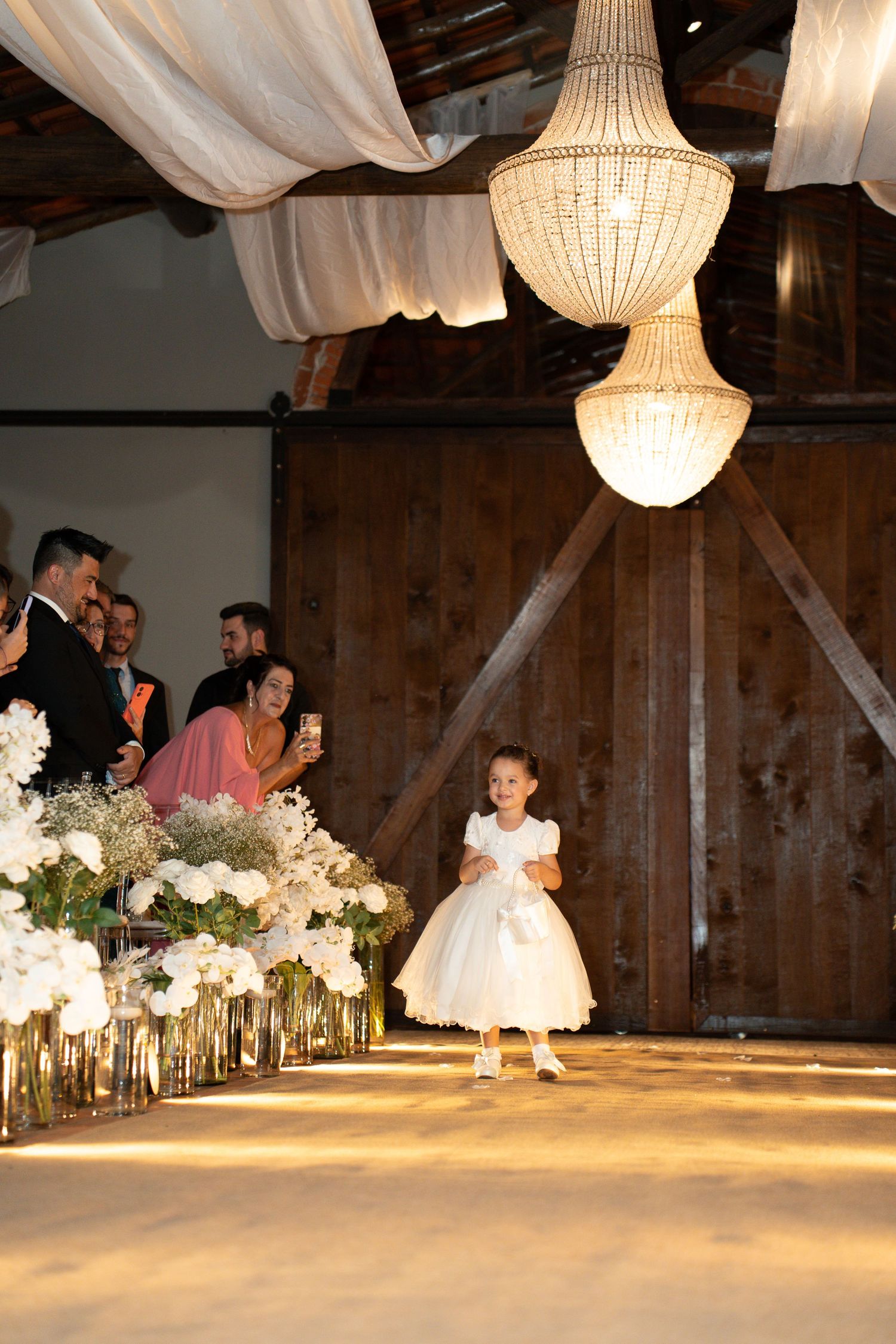 A flower girl in a white dress walks down a wedding aisle lined with white floral arrangements under elegant chandeliers.