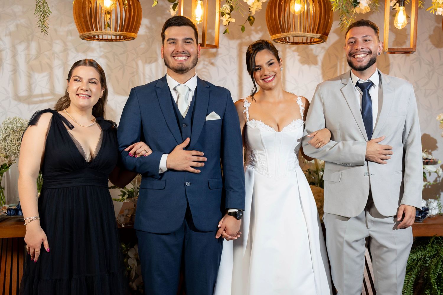 Bride and groom in wedding attire pose with two guests at elegant reception venue with warm lighting.