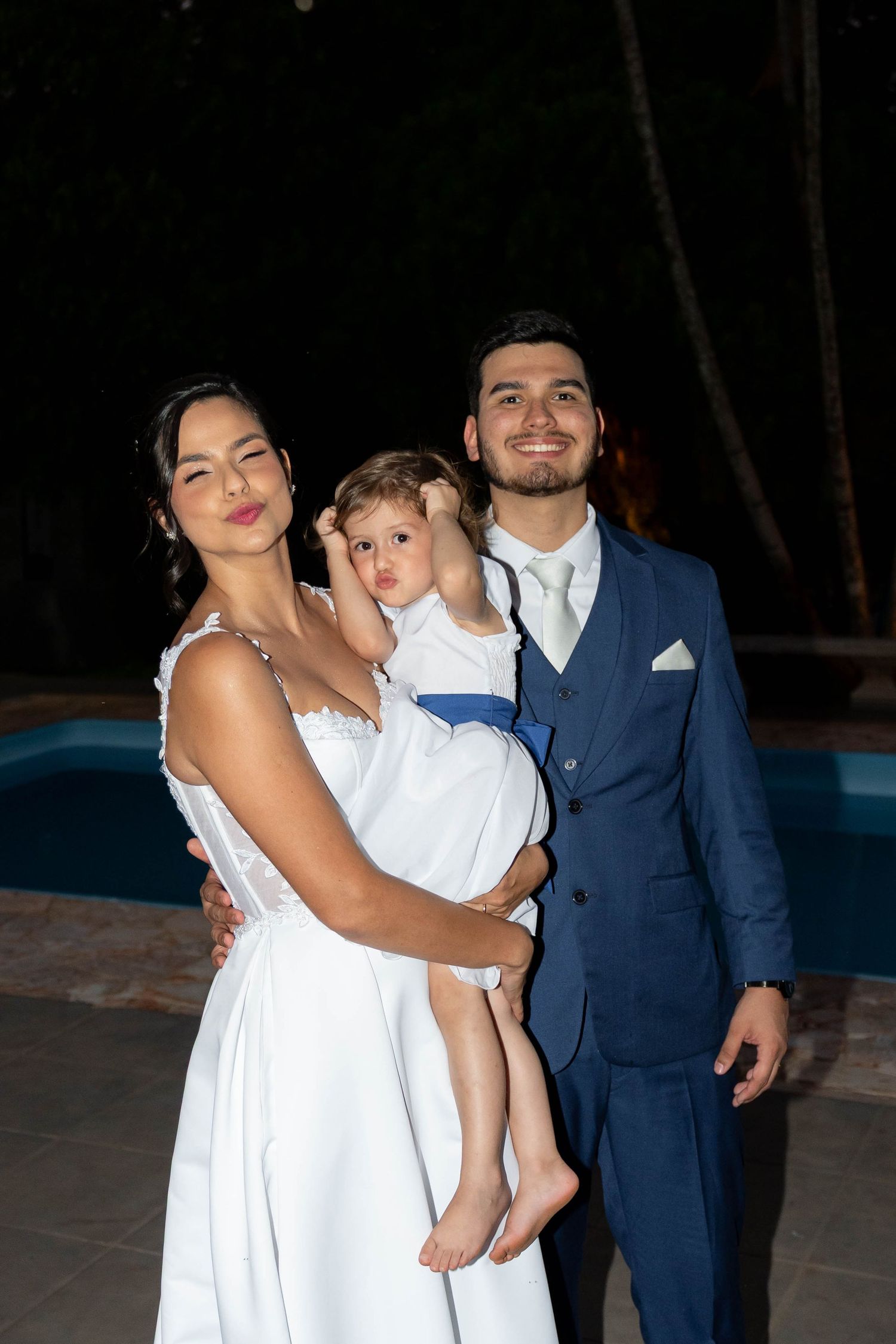 A smiling couple in wedding attire holds a young child in white near a pool at night.