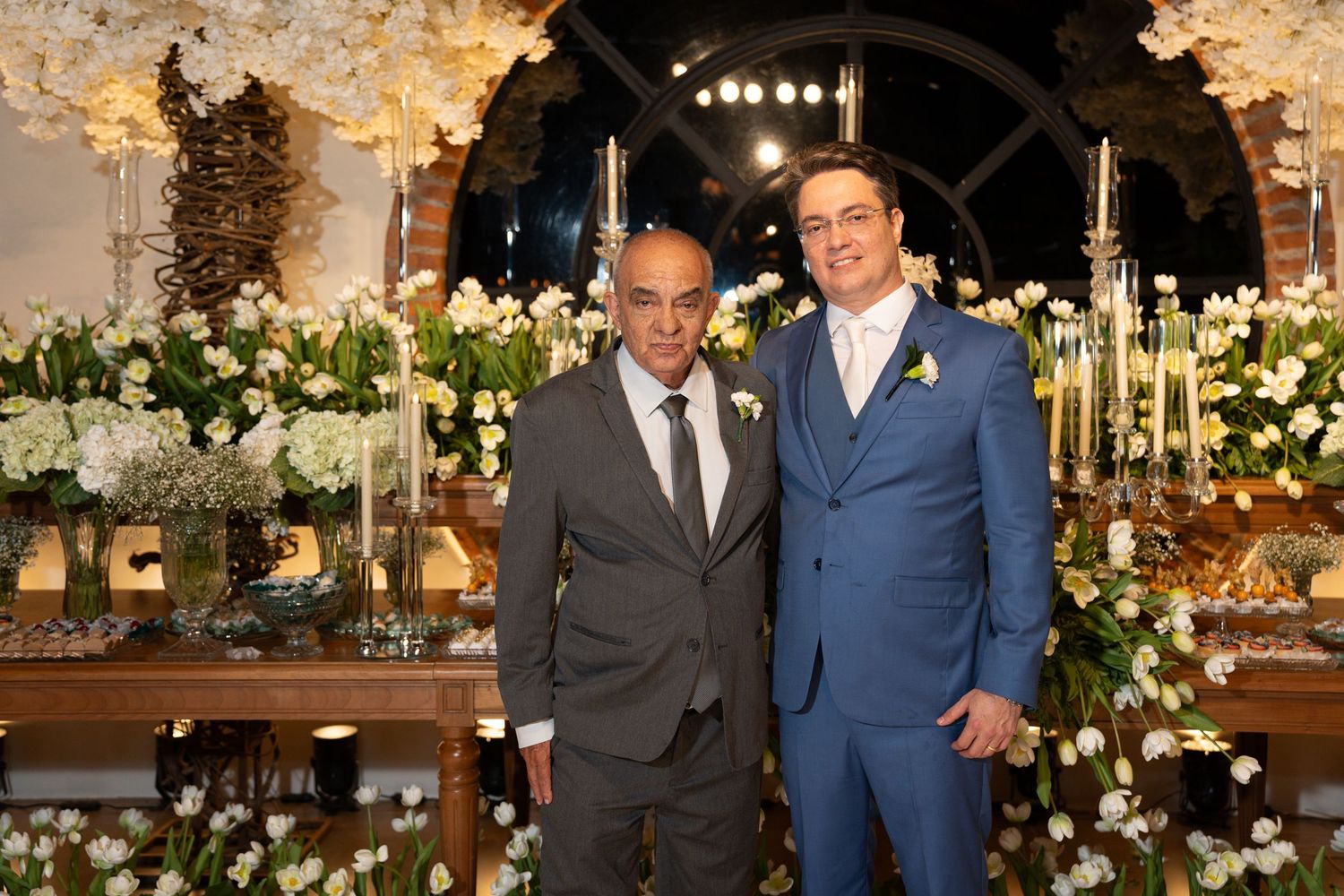 Two men in formal suits pose together at an elegant wedding reception with white floral arrangements and candelabras.