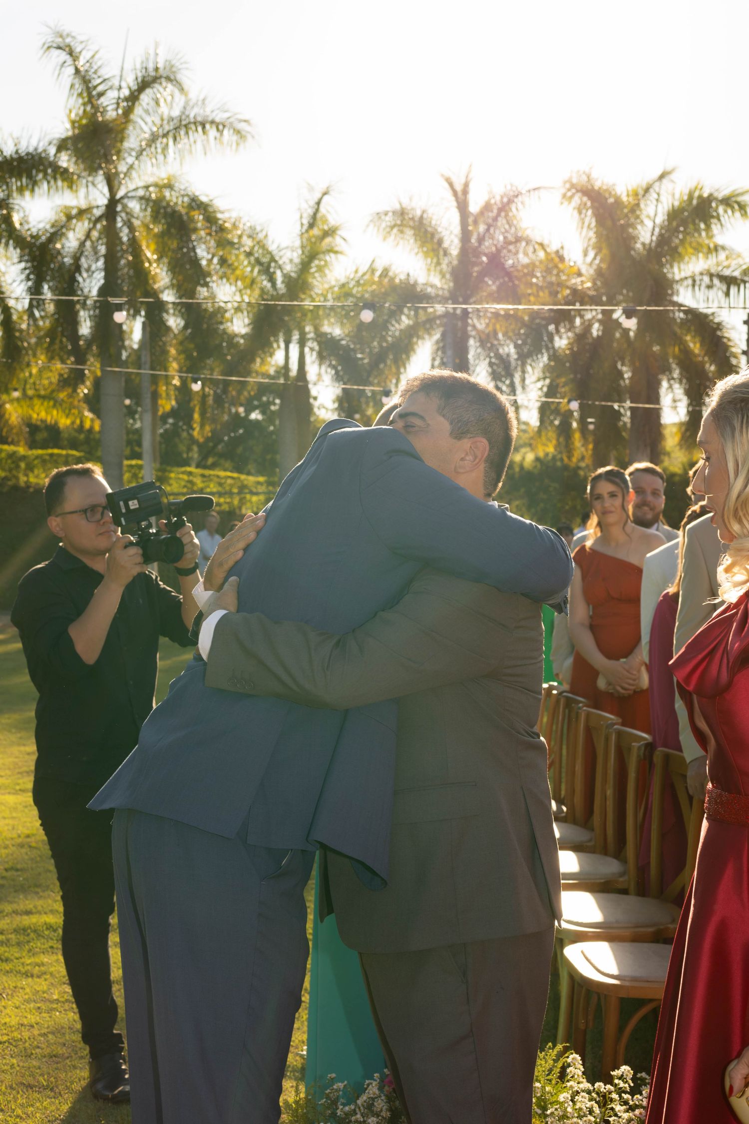 Two men share a warm hug at an outdoor wedding ceremony surrounded by palm trees and guests.