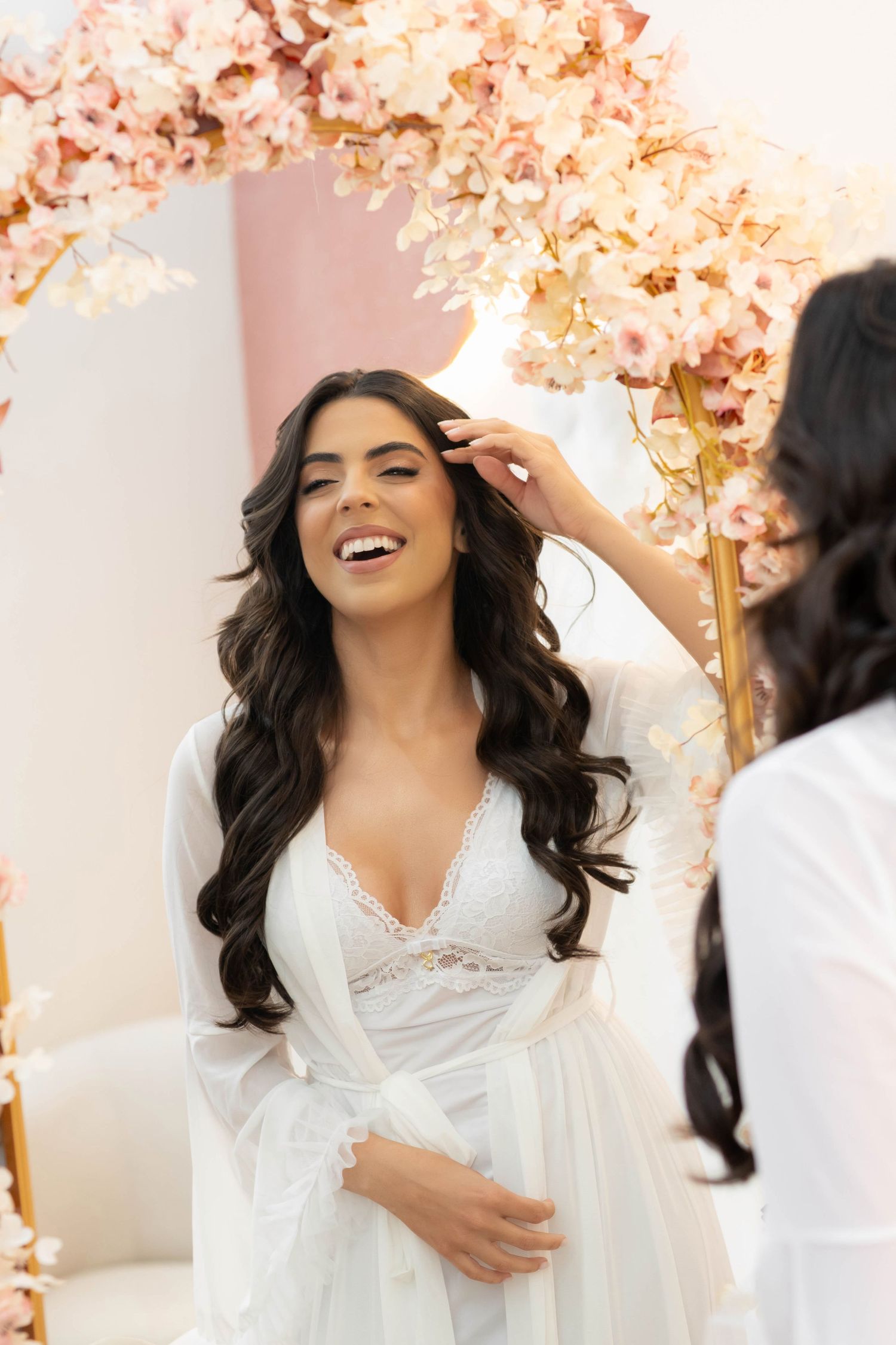 Smiling bride in white lace robe getting hair styled under pink floral arch during wedding preparation.