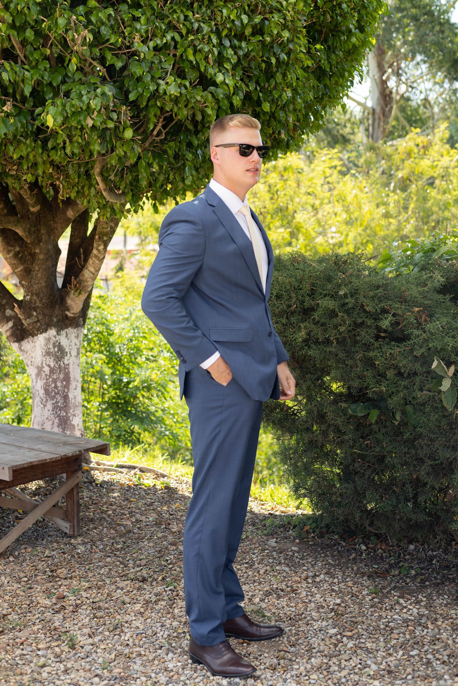 Man in blue suit and sunglasses standing outdoors in a garden with green trees and shrubs.