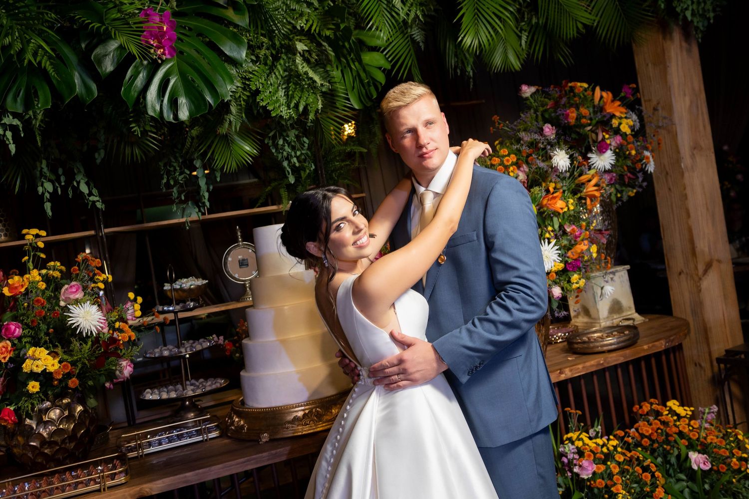 Bride and groom embrace in front of a floral wedding backdrop with a white tiered cake.