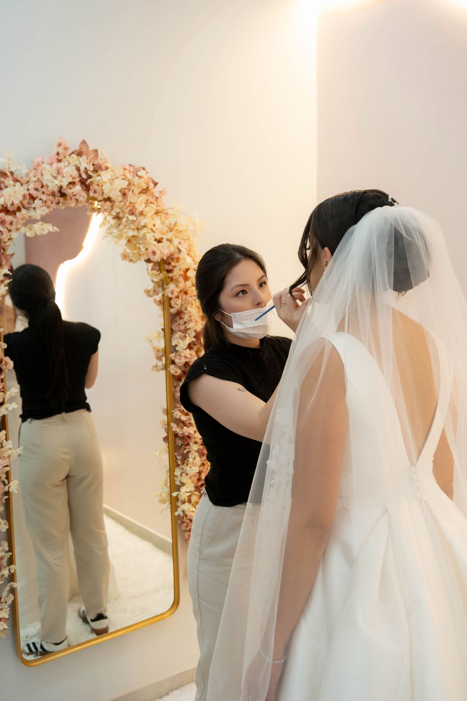 Makeup artist applying makeup to a bride in a white wedding dress and veil in front of a floral mirror.