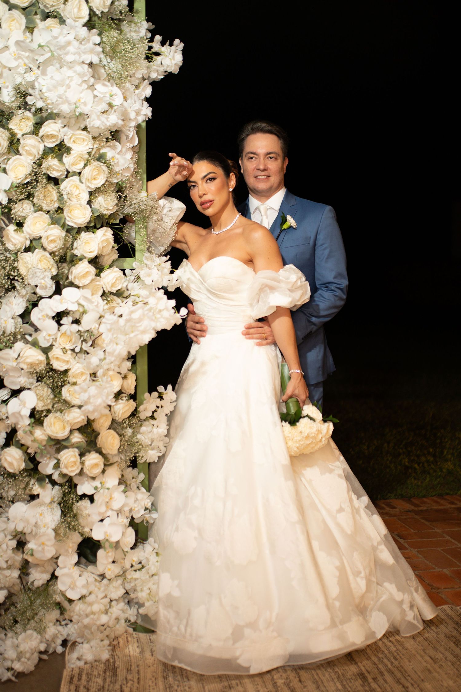 Elegant bride in strapless ball gown and groom in blue suit posing beside white floral arch at outdoor wedding ceremony.