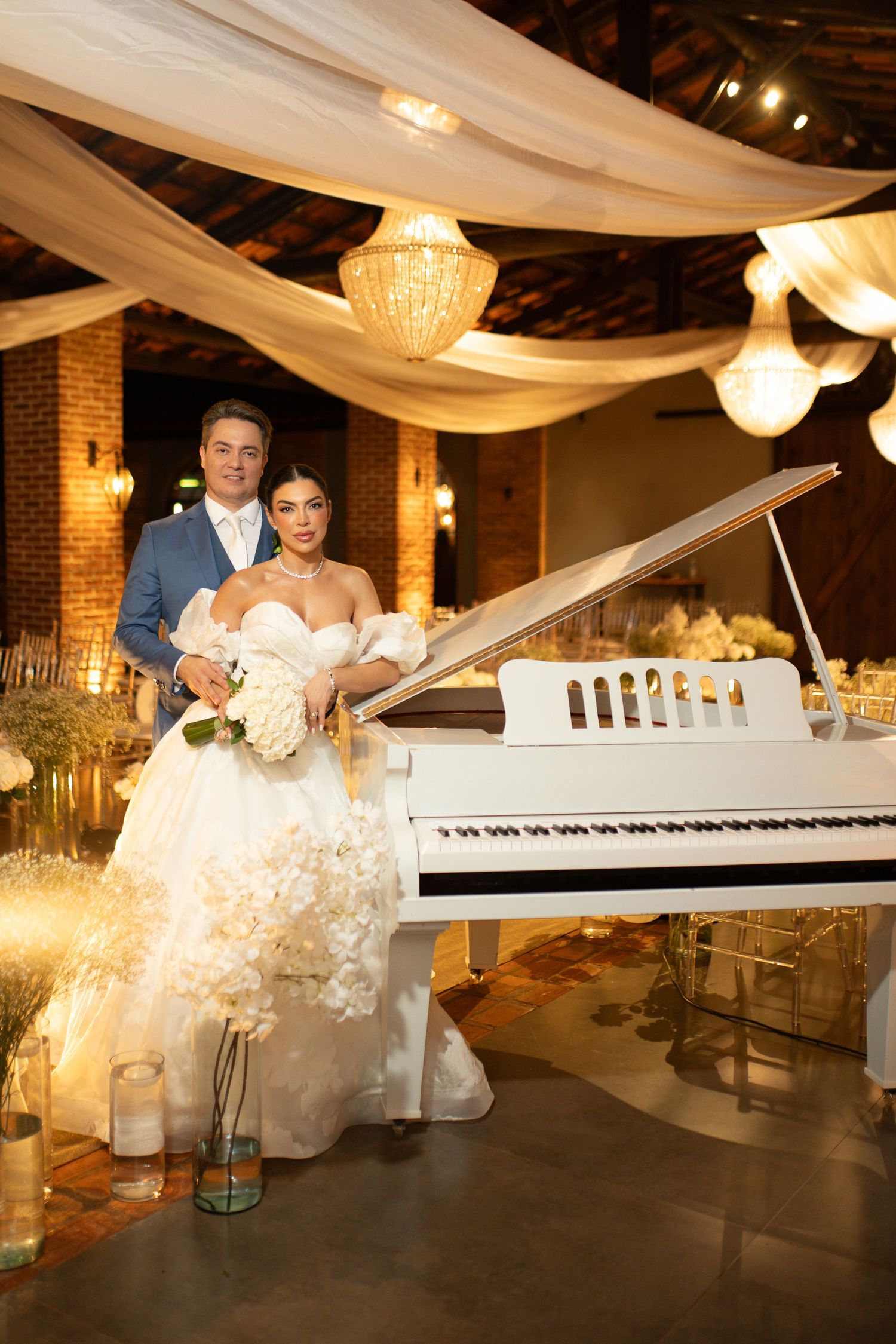Bride in white gown and groom in blue suit pose by a white grand piano at an elegant wedding reception.